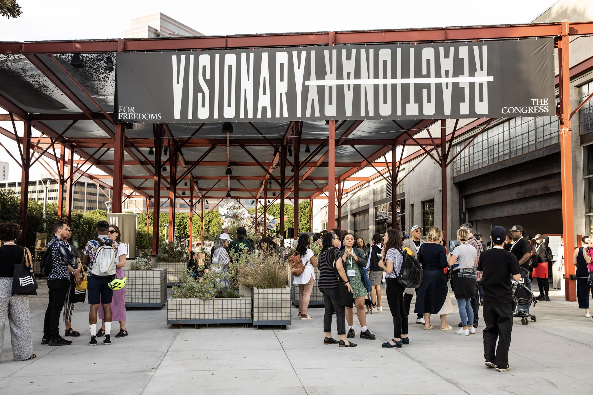 A diverse group of people gathered underneath a large outdoor canopy with a banner that reads "VISIONARY FUTURE" upside down. The setting appears to be an event or conference related to futures or innovation, with some individuals chatting, others st