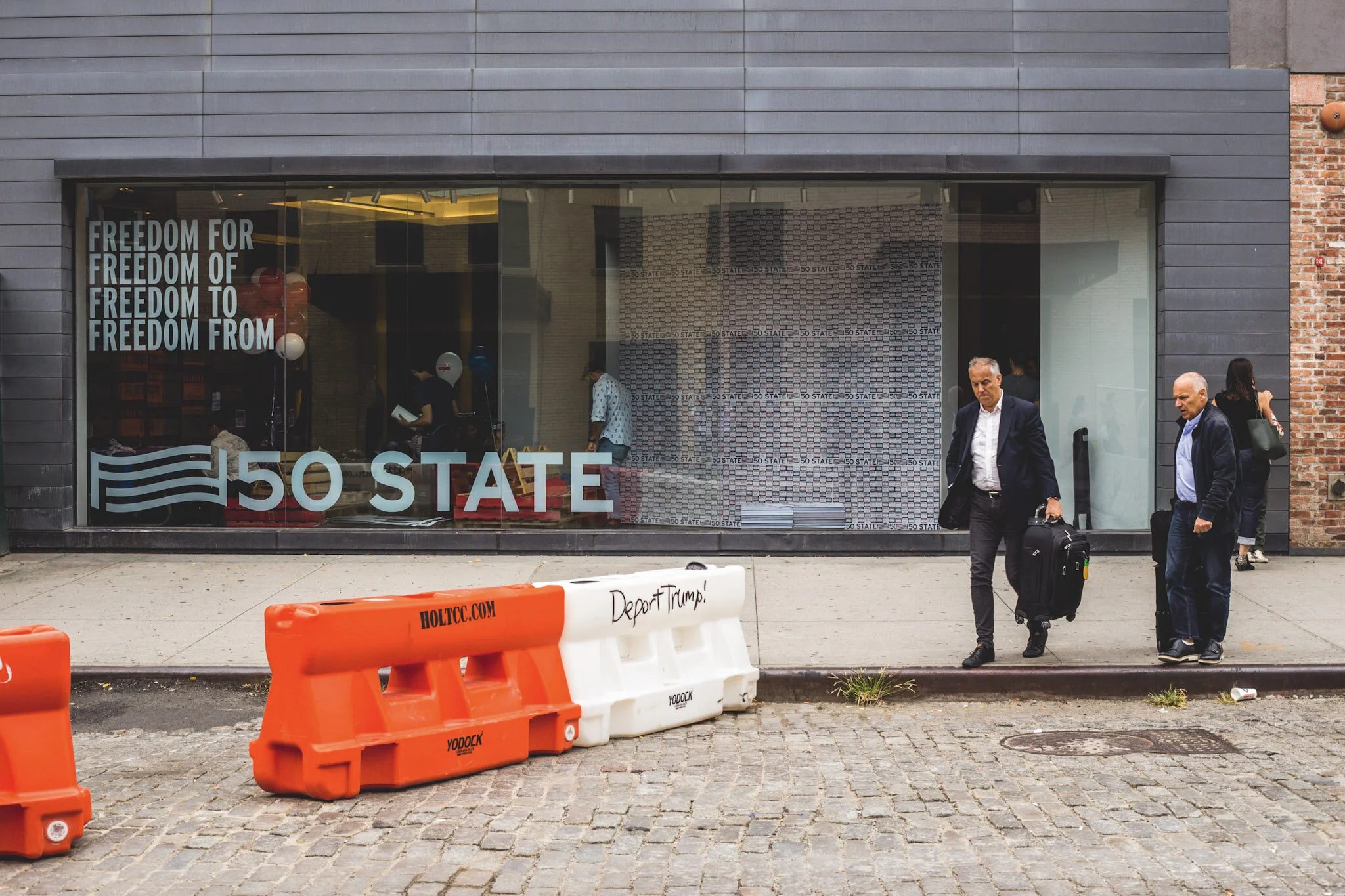 People walking past a storefront with '50 STATE' written on the window, including text that says 'FREEDOM FOR FREEDOM OF FREEDOM TO FREEDOM FROM'. In front, orange and white barricades with graffiti that reads 'Depart Trump!' and a website 'HOLTCC.CO
