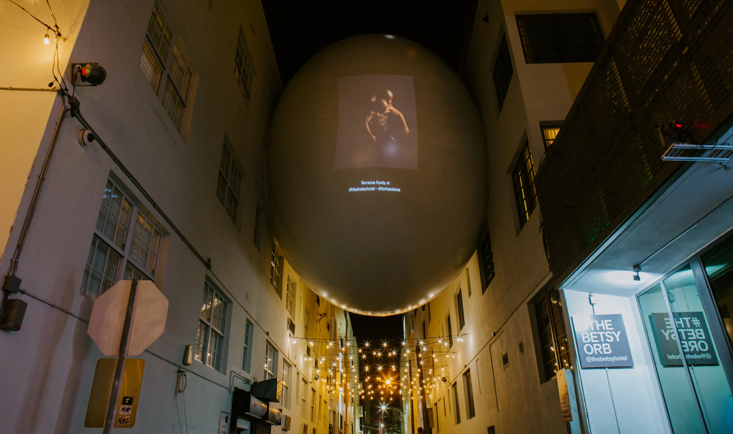 Night scene of an alleyway with hanging string lights, buildings on both sides, and a large spherical inflatable structure with a projected image of a woman on it.
