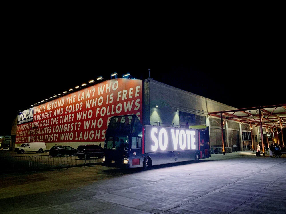 Nighttime scene outside a large building with a billboard and a mobile voting trailer. The billboard displays questions about law, ownership, and existence. The trailer has a bright illuminated sign that reads 'SO VOTE'.