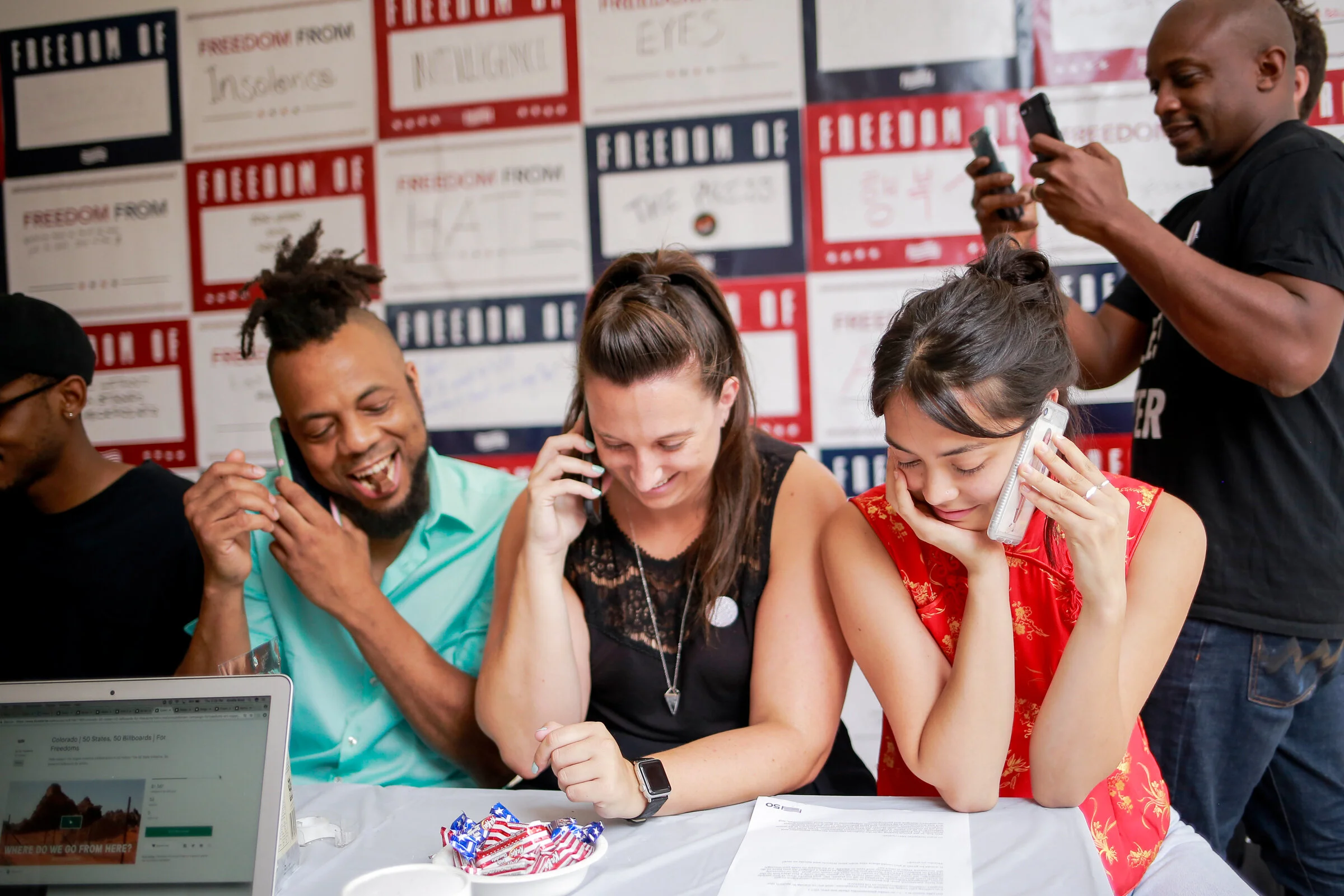 Group of four diverse people sitting at a table, smiling and talking on phones, with a man standing behind taking a photo, in front of a wall decorated with patriotic posters related to freedom and rights.