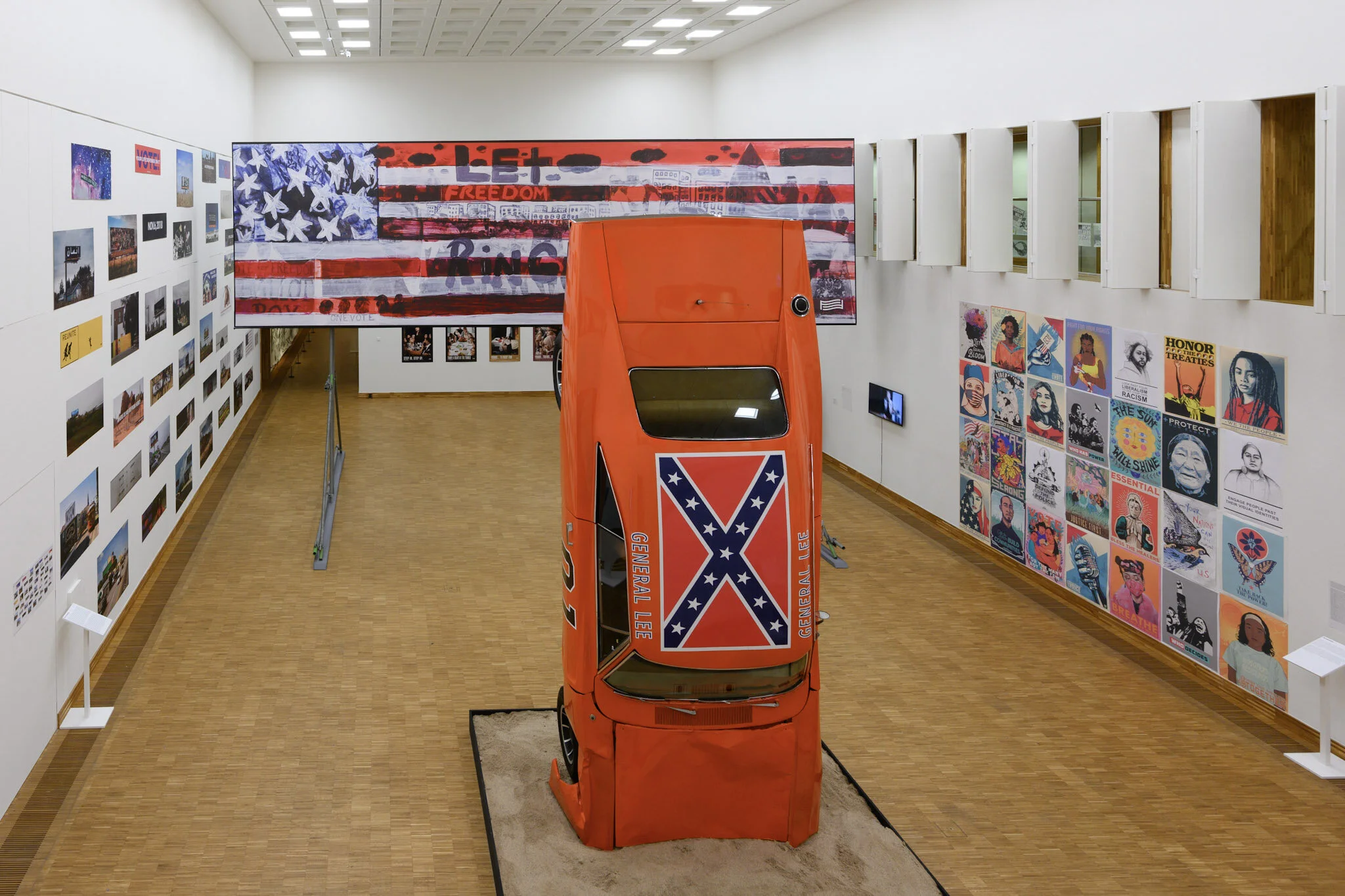 An art exhibit with a red vehicle featuring a Confederate flag painted on it, displayed upside down inside a gallery. The walls are decorated with various artwork and photographs, and there is a large American flag and slogans painted on a panel in t