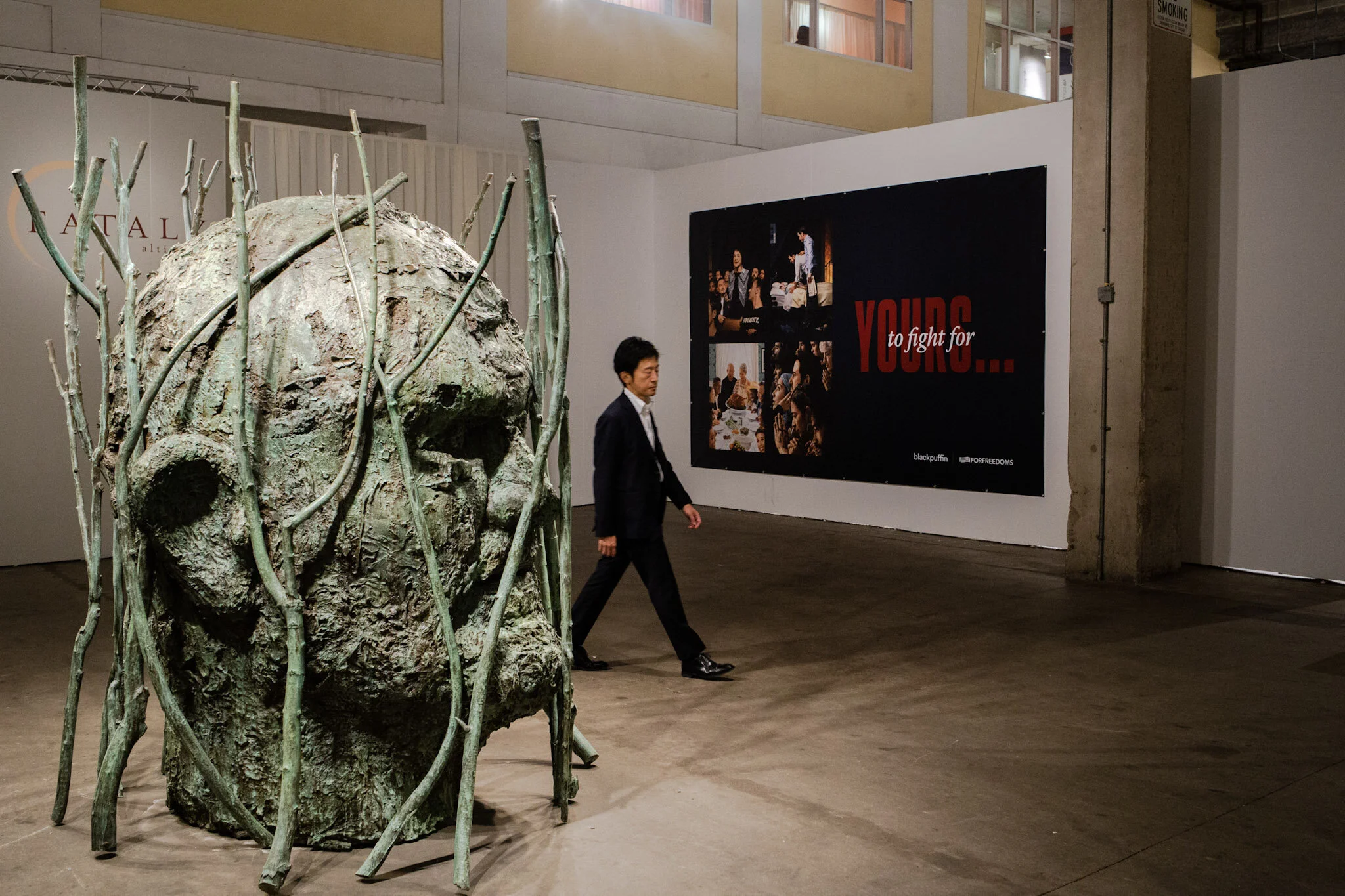 An art installation resembling a large, weathered human face with hollow eyes and surrounded by thick, stringy vegetation in an indoor gallery space. A man in a black suit walks past it, with large posters on the white walls behind him.