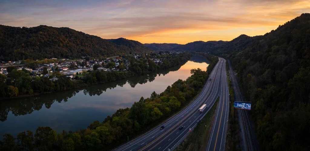 An aerial view of a winding highway running through a valley surrounded by forested hills during sunset, with a calm river reflecting the sky.
