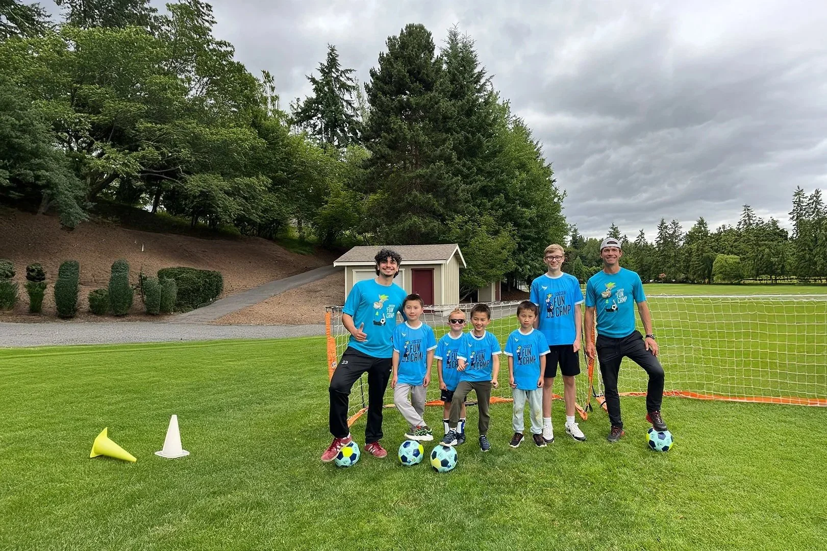 Group of children and two coaches standing on a soccer field with soccer balls, goal net, and cones, during a soccer camp, with trees and overcast sky in the background.