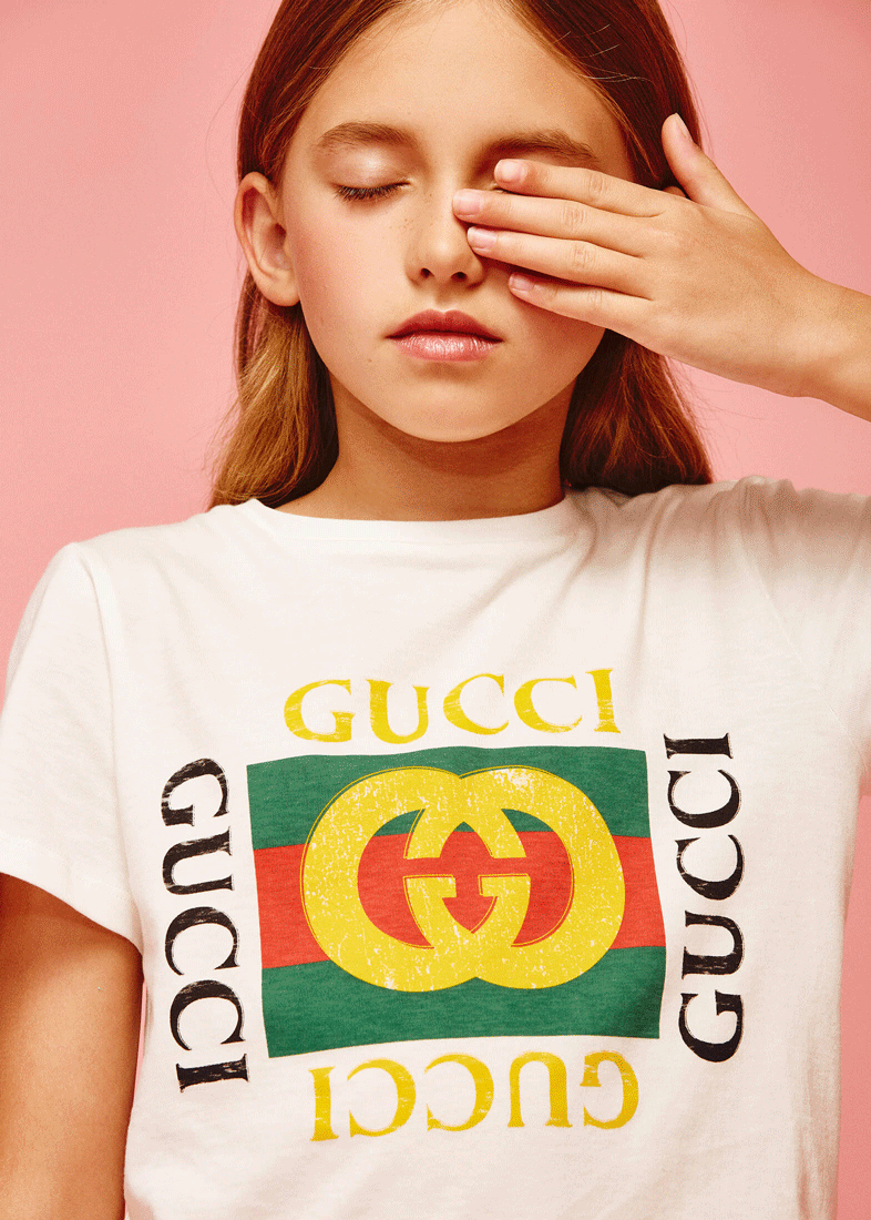 Children’s fashion photography in studio by kids fashion photographer Vika Pobeda captures a bright and cheerful portrait of a young girl wearing a stylish Gucci T-shirt. Her playful pose, with one hand partially covering her face, highlights her rad