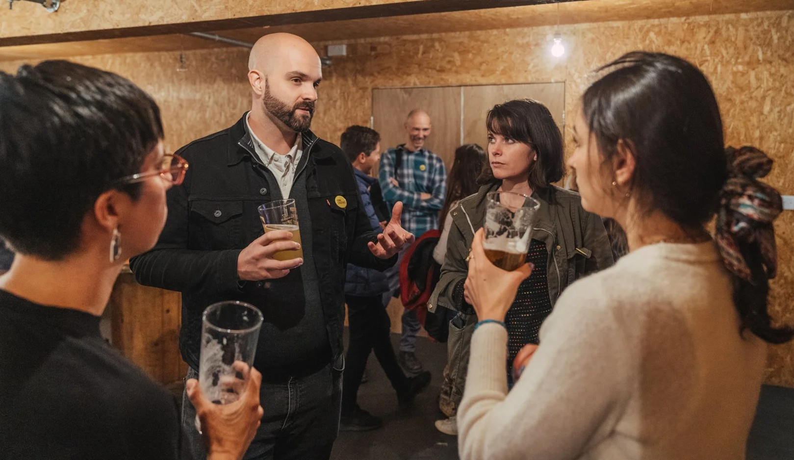 Group of four people having a conversation at a social gathering with wooden walls, holding drinks, in a casual bar or brewery environment.