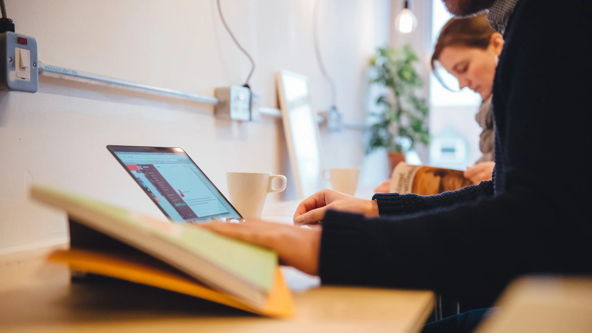 Person working on a laptop at a wooden table in a cafe or workspace, with books and a coffee mug nearby, and another person in the background reading a book.