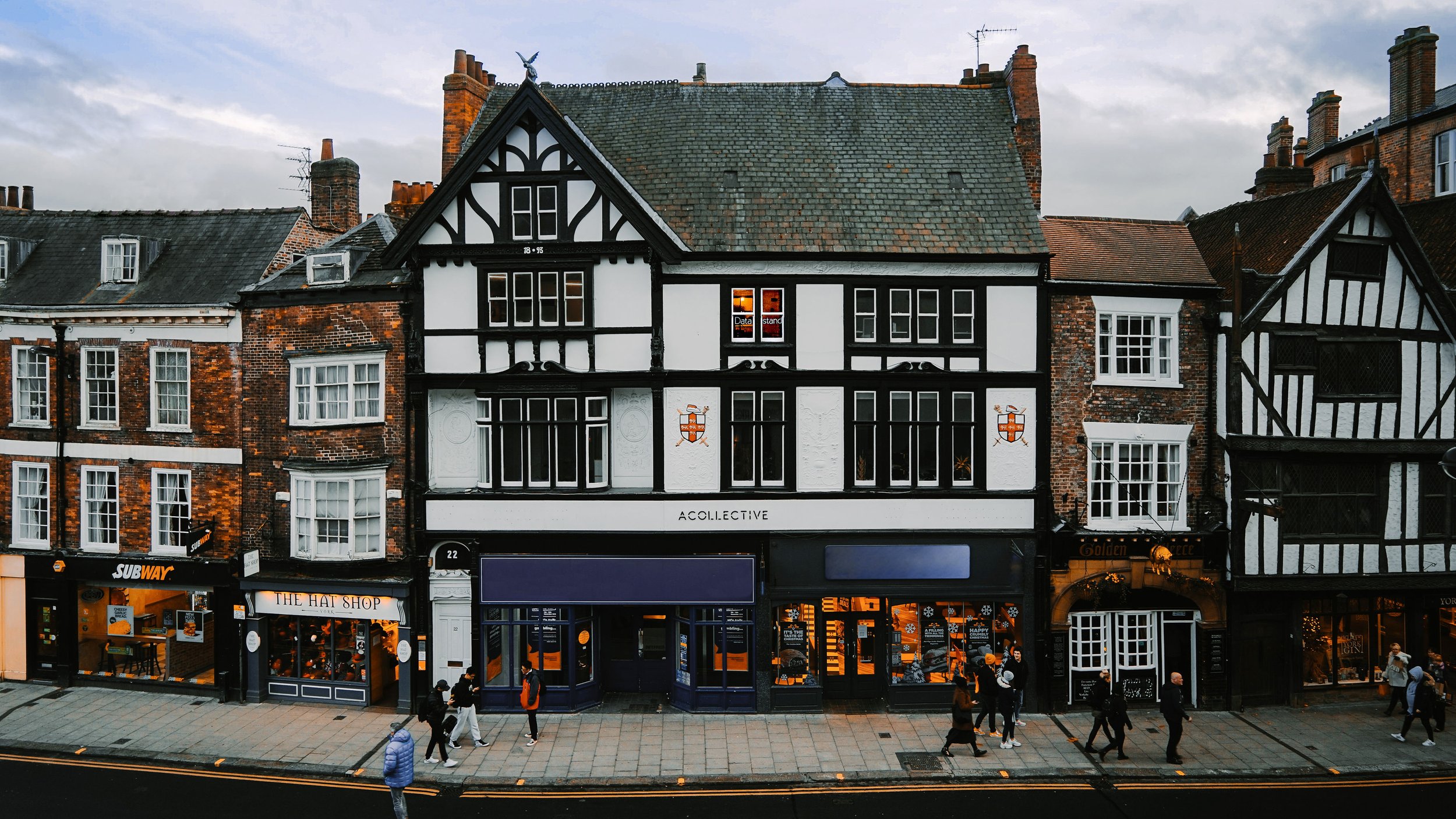 Street view showing a row of charming historical buildings with shops on the ground floor, including Subway, The Hat Shop, and a pub called Golden Ace, with people walking along the sidewalk in front.