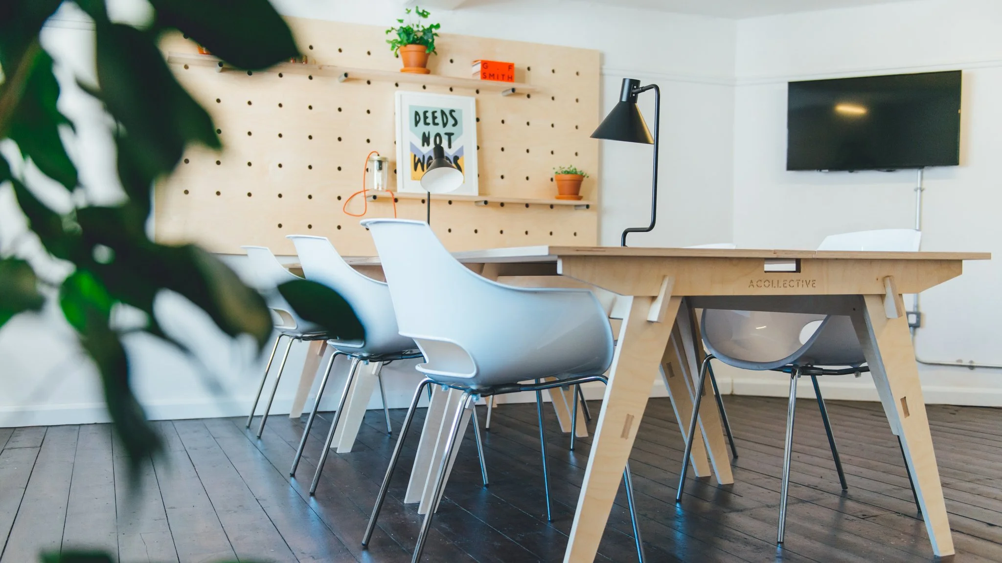 Modern conference room with wooden table, white chairs, pegboard wall with framed art, potted plants, hanging lights, and a wall-mounted television.