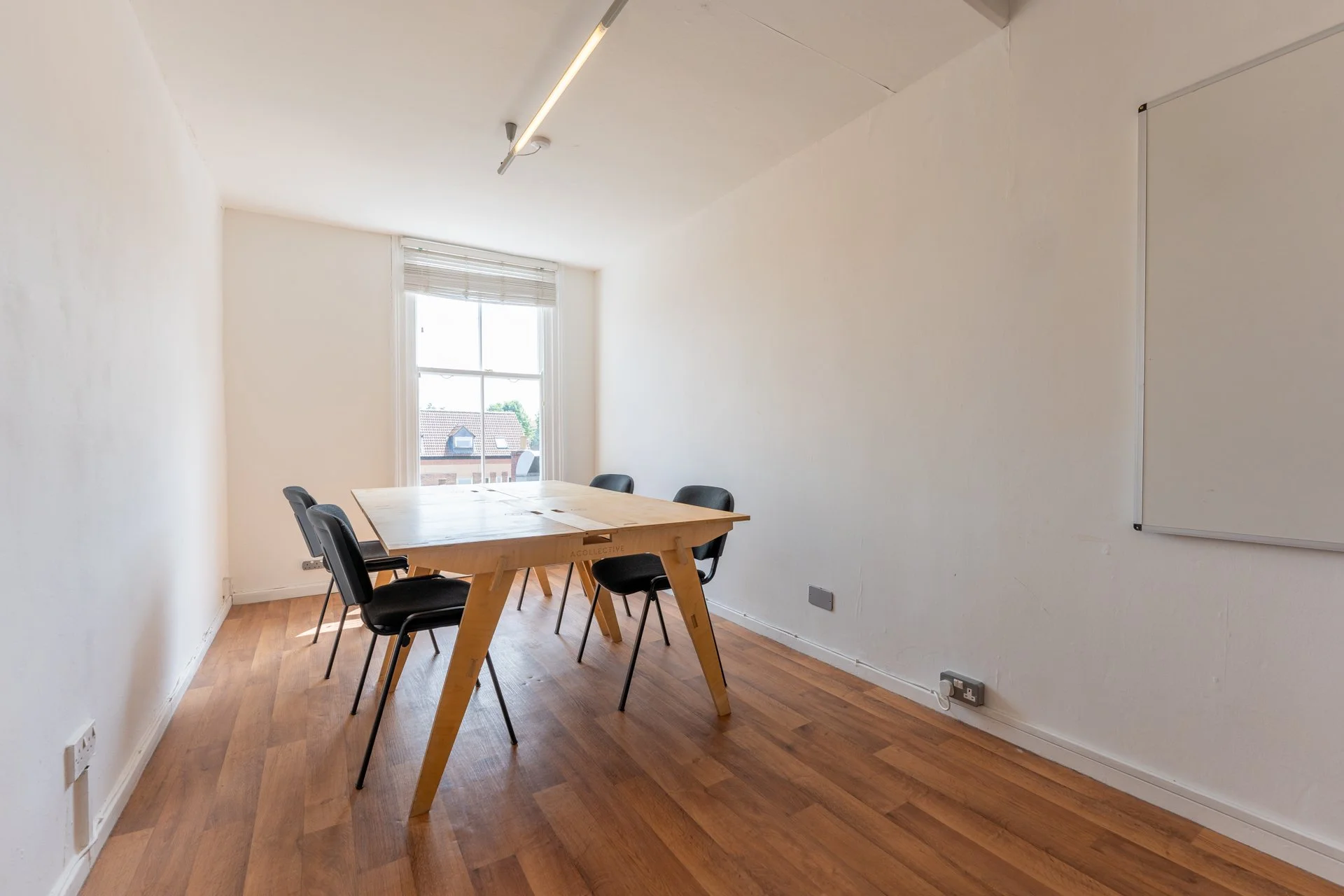 Empty meeting room with a wooden table, four black chairs, a window with blinds, and a whiteboard on the right wall.