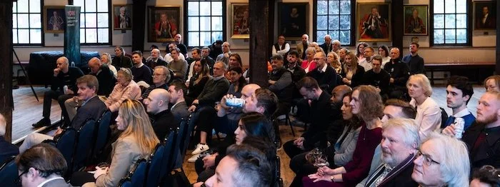 An audience seated in a conference room with paintings on the walls, attending a presentation or lecture.