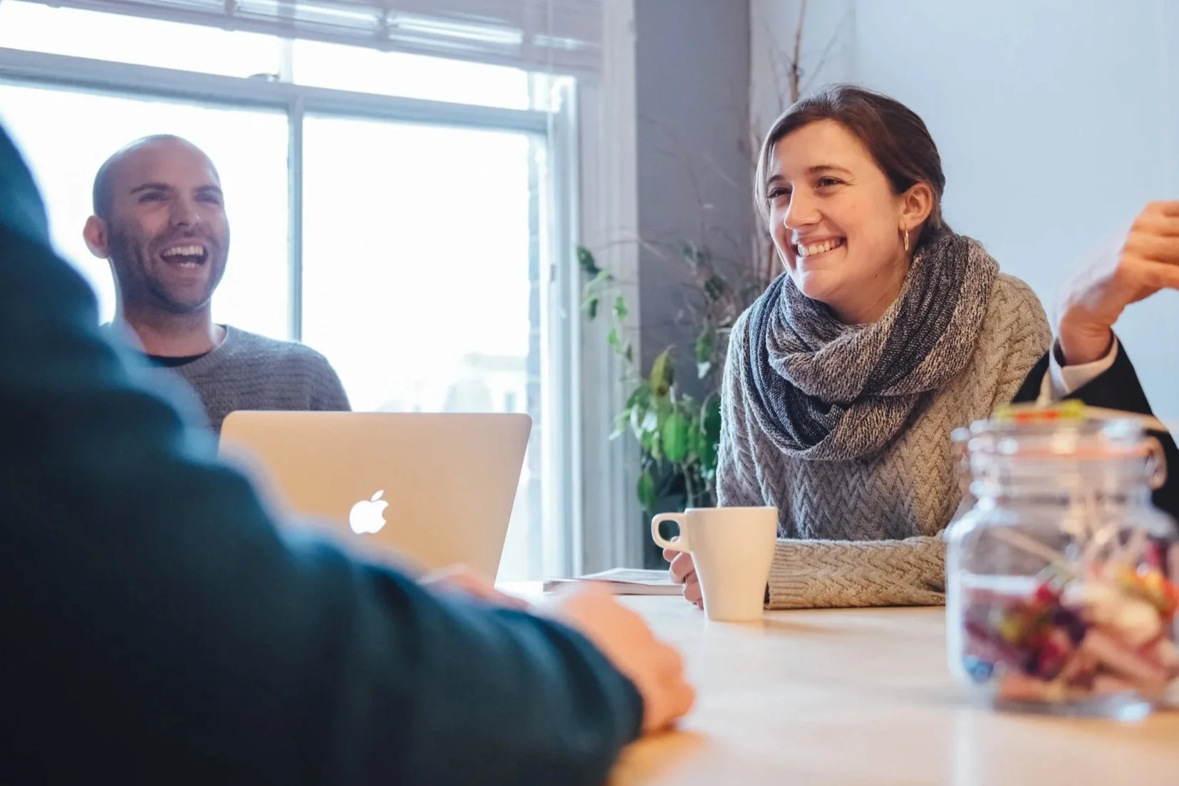 Three people sitting at a table having a conversation, smiling, with a laptop, a mug, and a jar of candy on the table, in a bright room with large windows and plants.