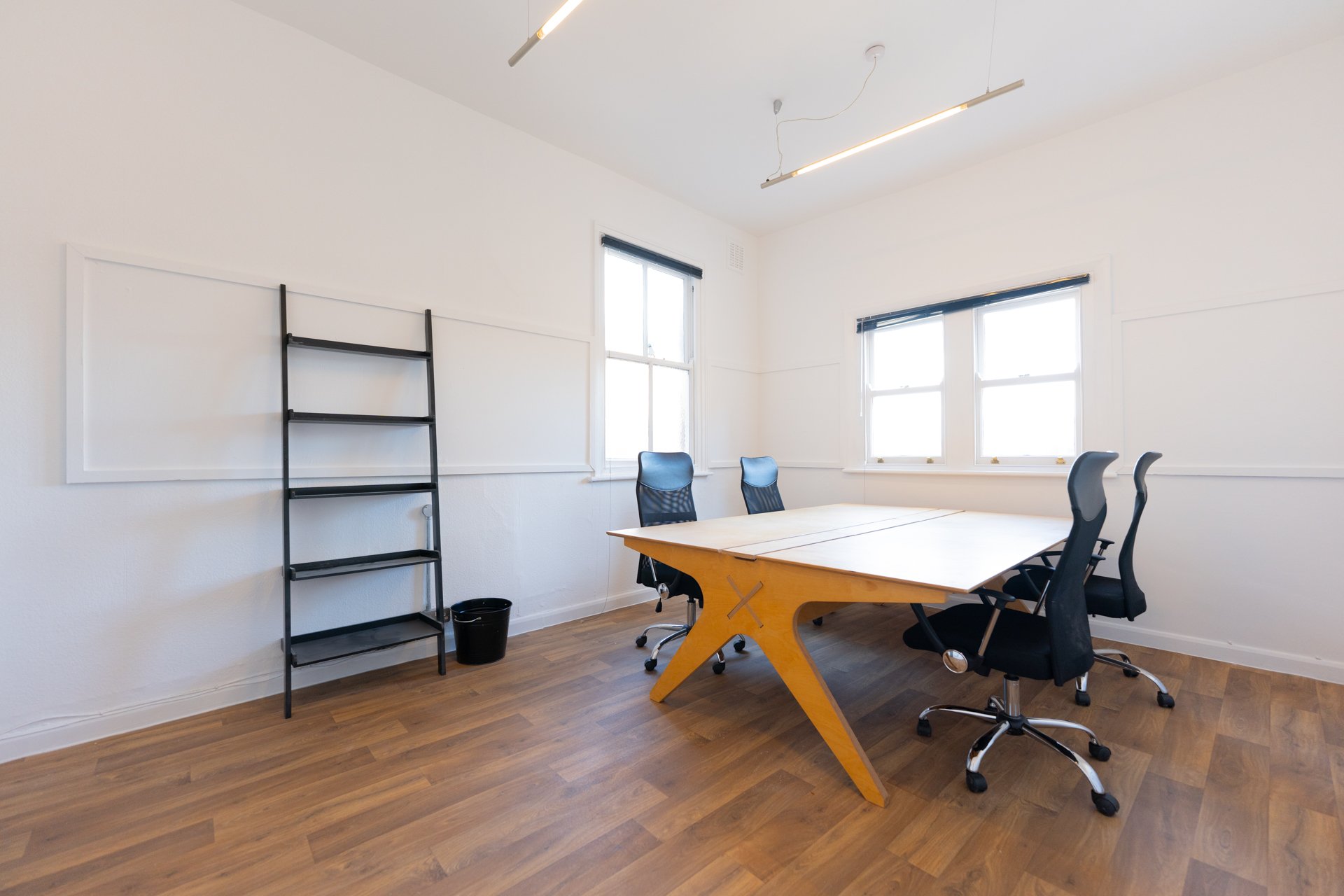 A conference room with a large wooden table, black office chairs, white walls, two large windows, and a black metal ladder shelf with shelves and a small black trash bin underneath.