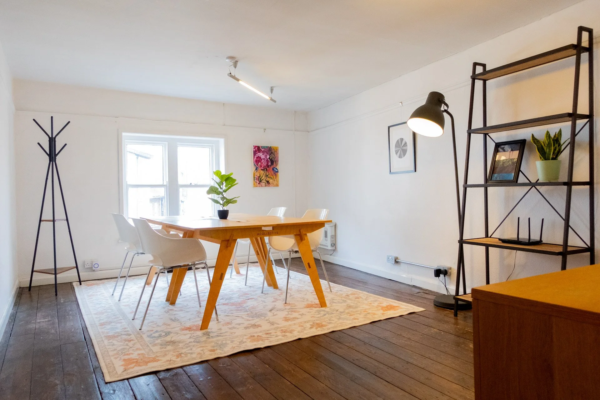 Bright dining room with a wooden table, white chairs, and a plant in a black pot. There is a window with natural light, a colorful art piece on the wall, and a black metal shelf with framed pictures and a green plant. A coat rack and a standing lamp are also visible.