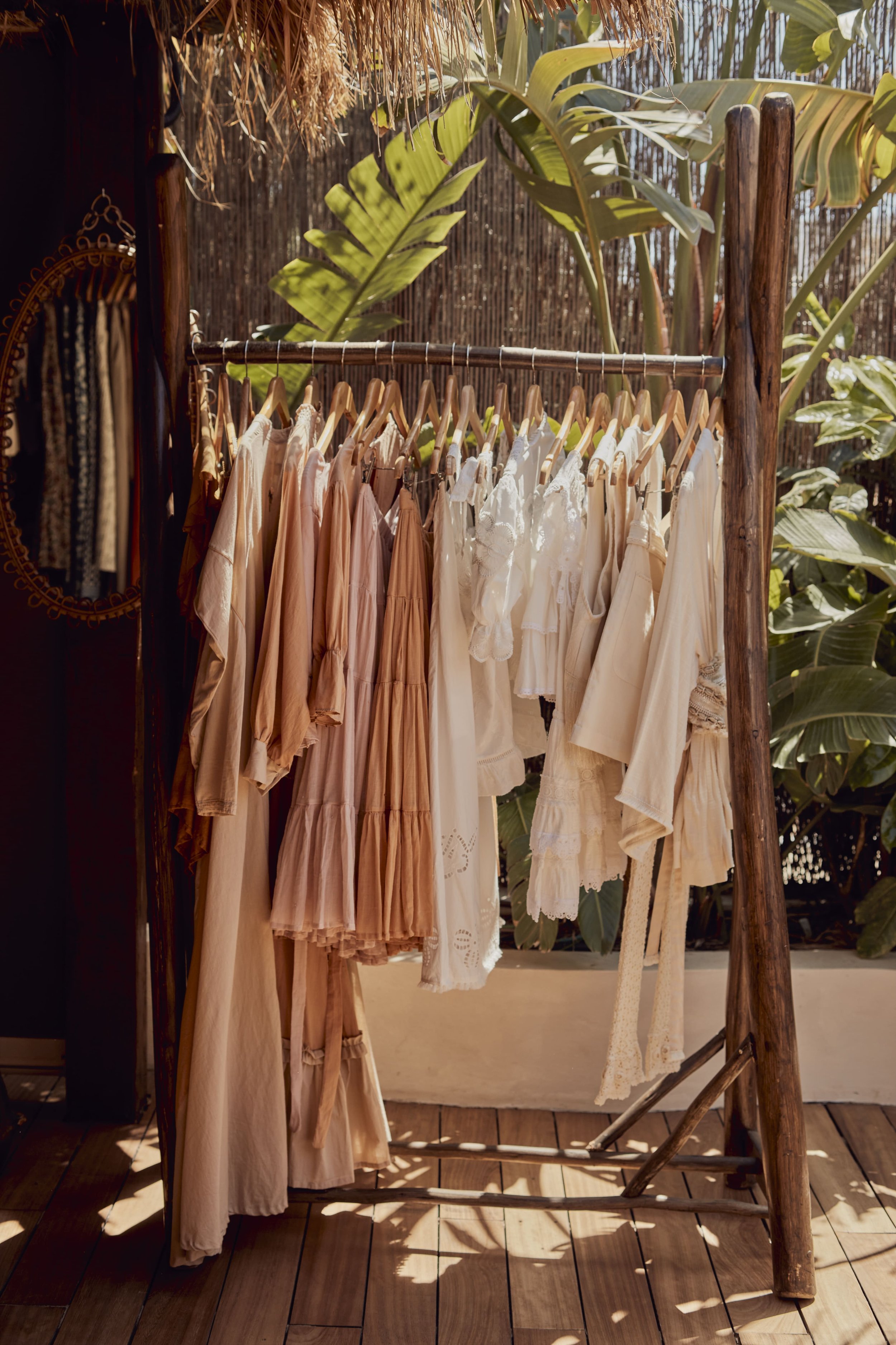 Clothing rack with beige and white dresses and tops outdoors, surrounded by leafy plants and bamboo fencing.