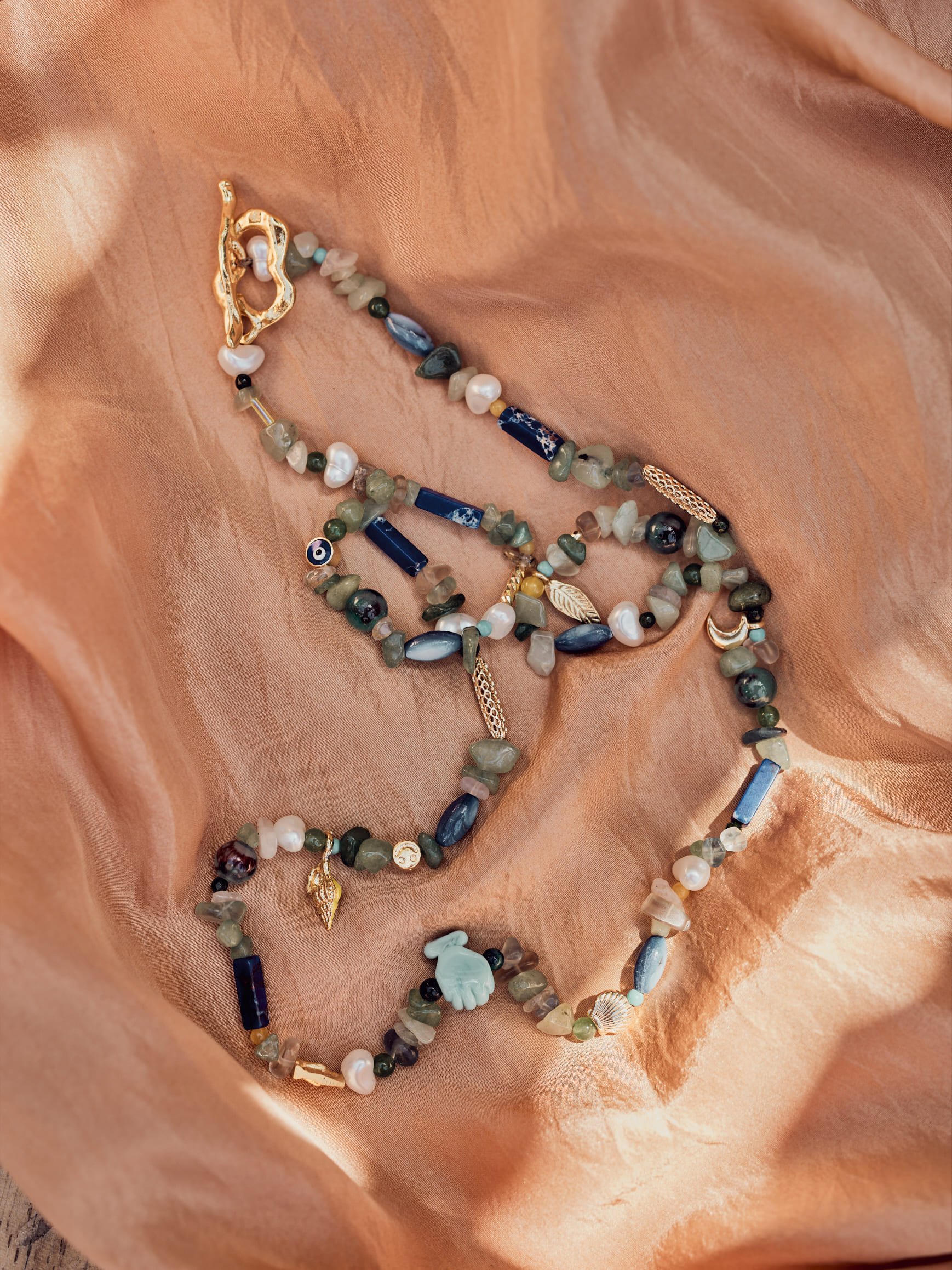 A beaded necklace with various shells, stones, and charms, including gold-colored leaf and hand charms, resting on peach-colored satin fabric.