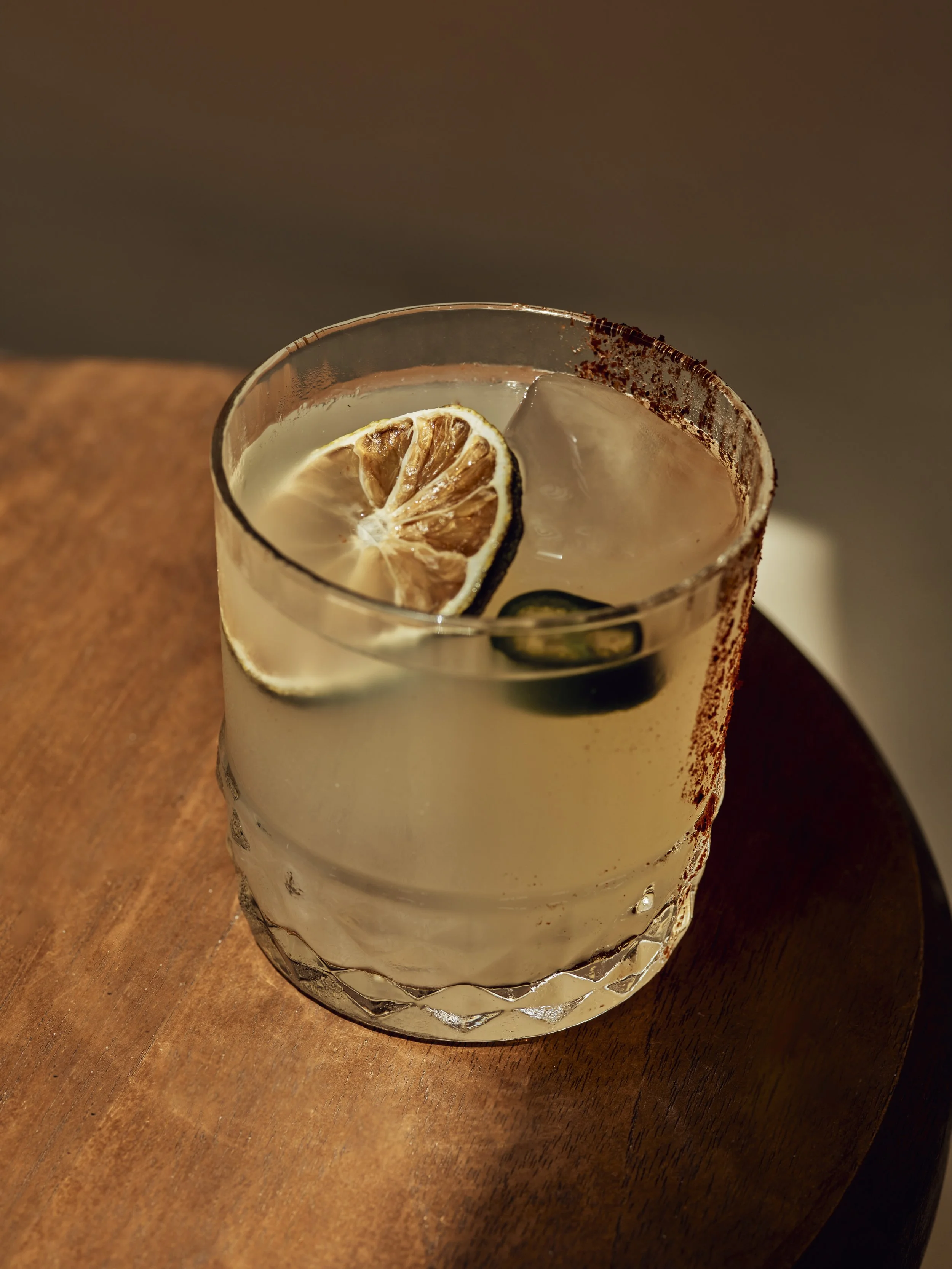 A glass of lemonade with slices of lemon and cucumber, rimmed with salt and chili powder, placed on a wooden table.