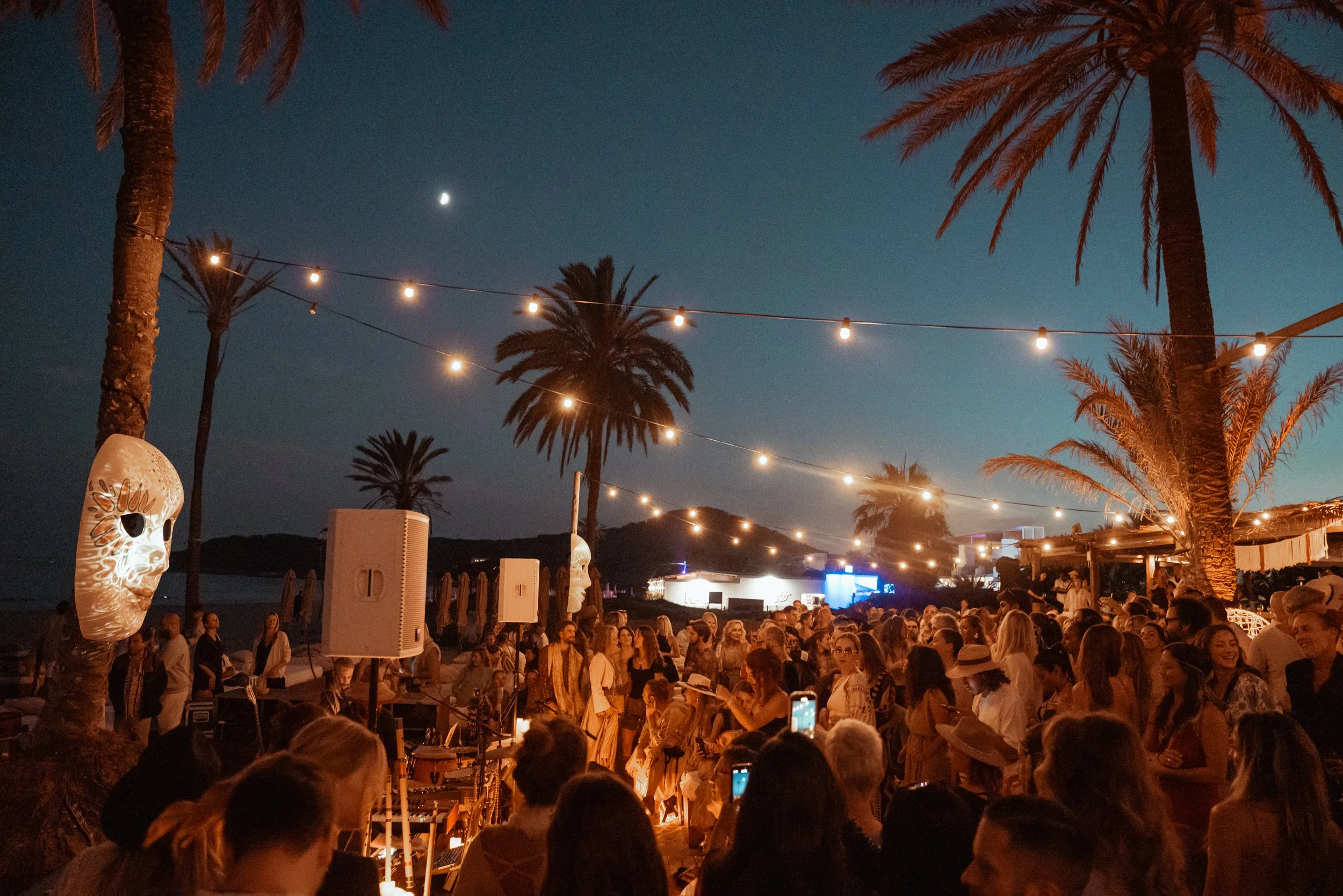 A large crowd gathered outdoors at night under string lights. Tall palm trees are visible, with a crescent moon in the sky. There are also white decorative lanterns among the crowd, and some people are taking photos or videos with their phones.