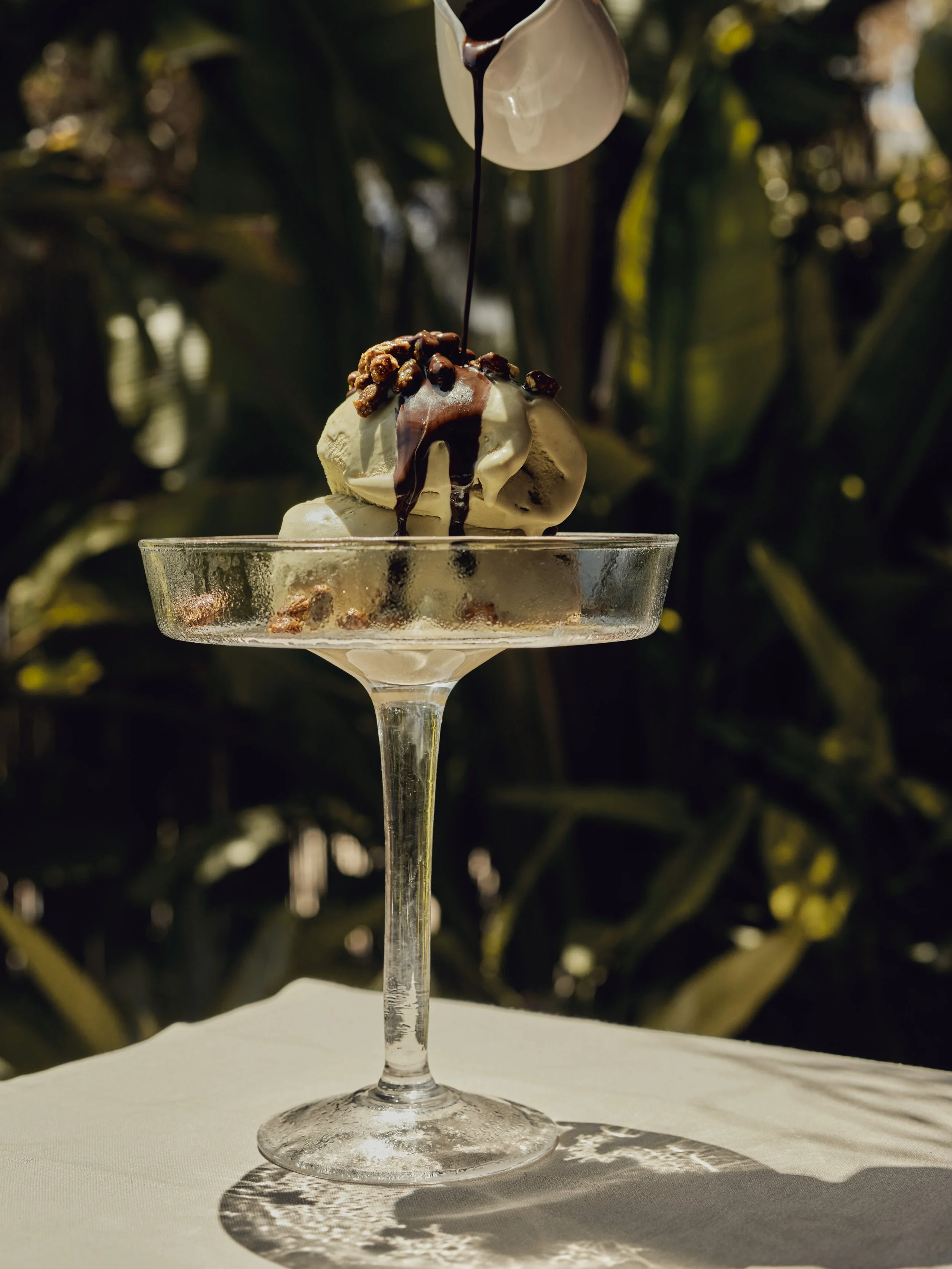 A glass dessert dish with a scoop of vanilla ice cream topped with chocolate syrup and caramelized nuts. Chocolate syrup is being poured over the ice cream from a white pitcher. Sunlight creates a pattern of shadows on the surface of the dish, with a