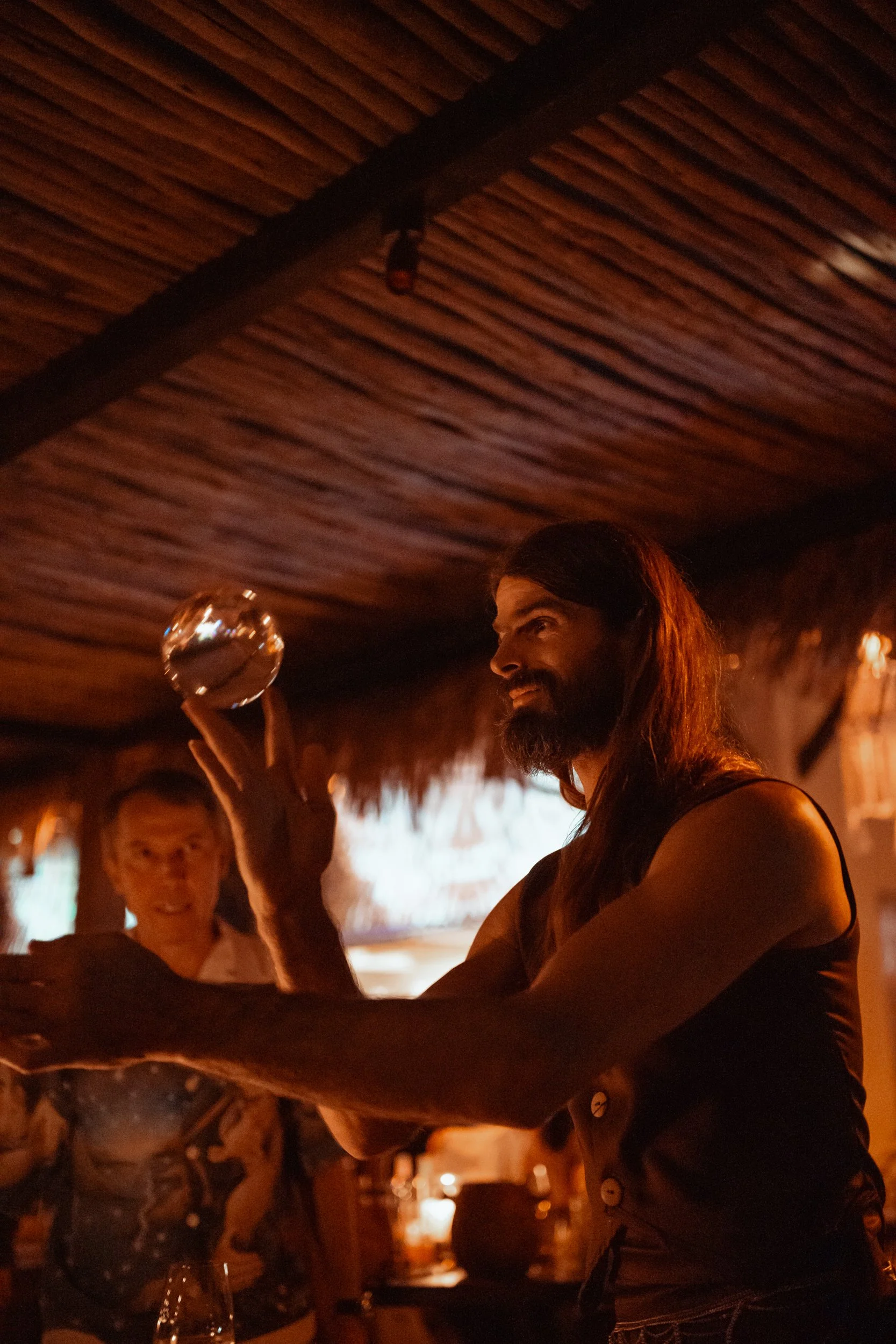 A group of people at a bar or restaurant enjoying drinks and conversation in a cozy, dimly lit setting with a tropical or tiki theme, featuring a wooden ceiling and warm lighting.