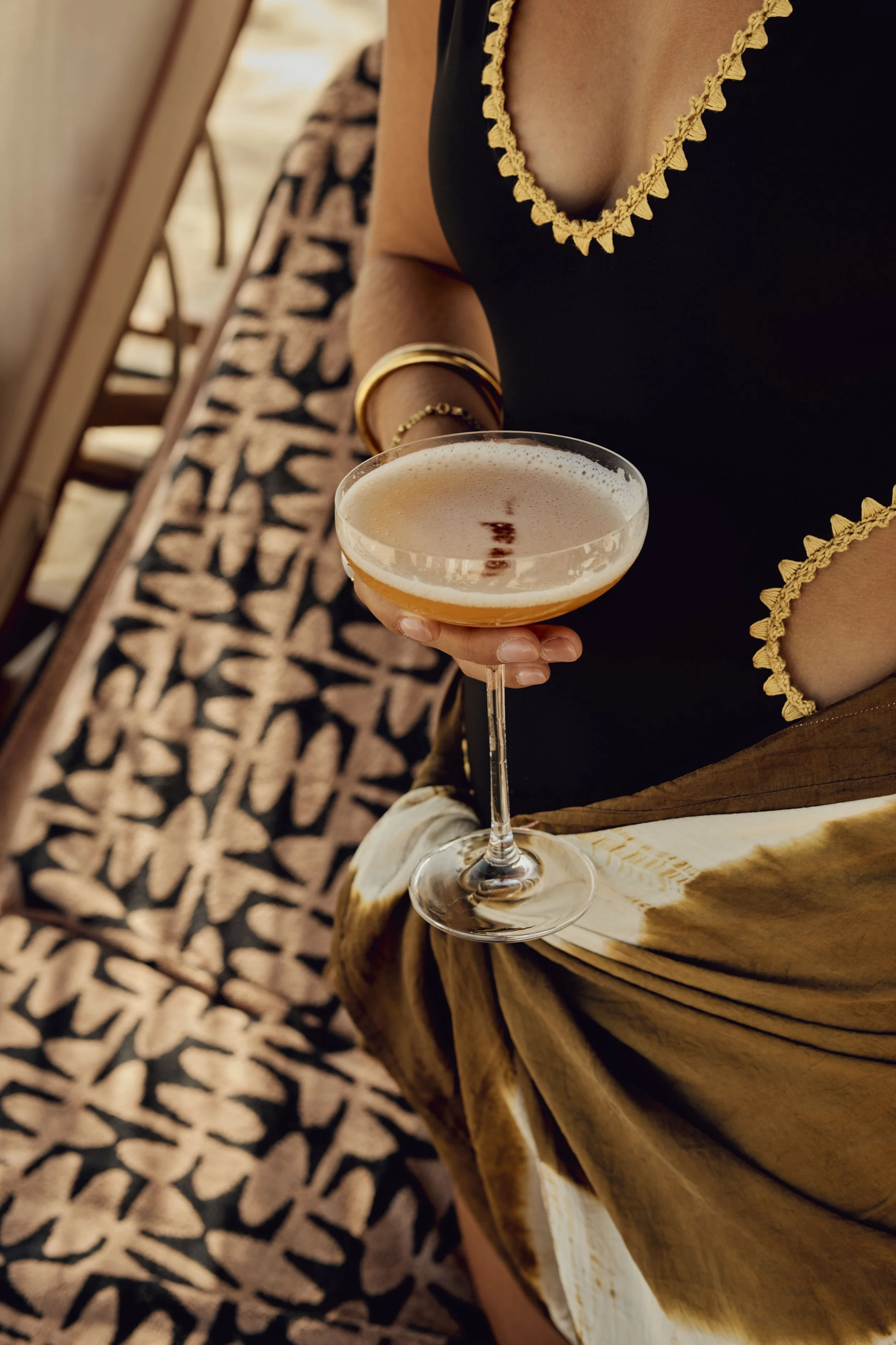 A woman wearing a black top with yellow trim holds a cocktail glass with a pinkish drink and frothy top, dressed with a gold bracelet and ring, sitting at a wooden table with a patterned fabric in the background.
