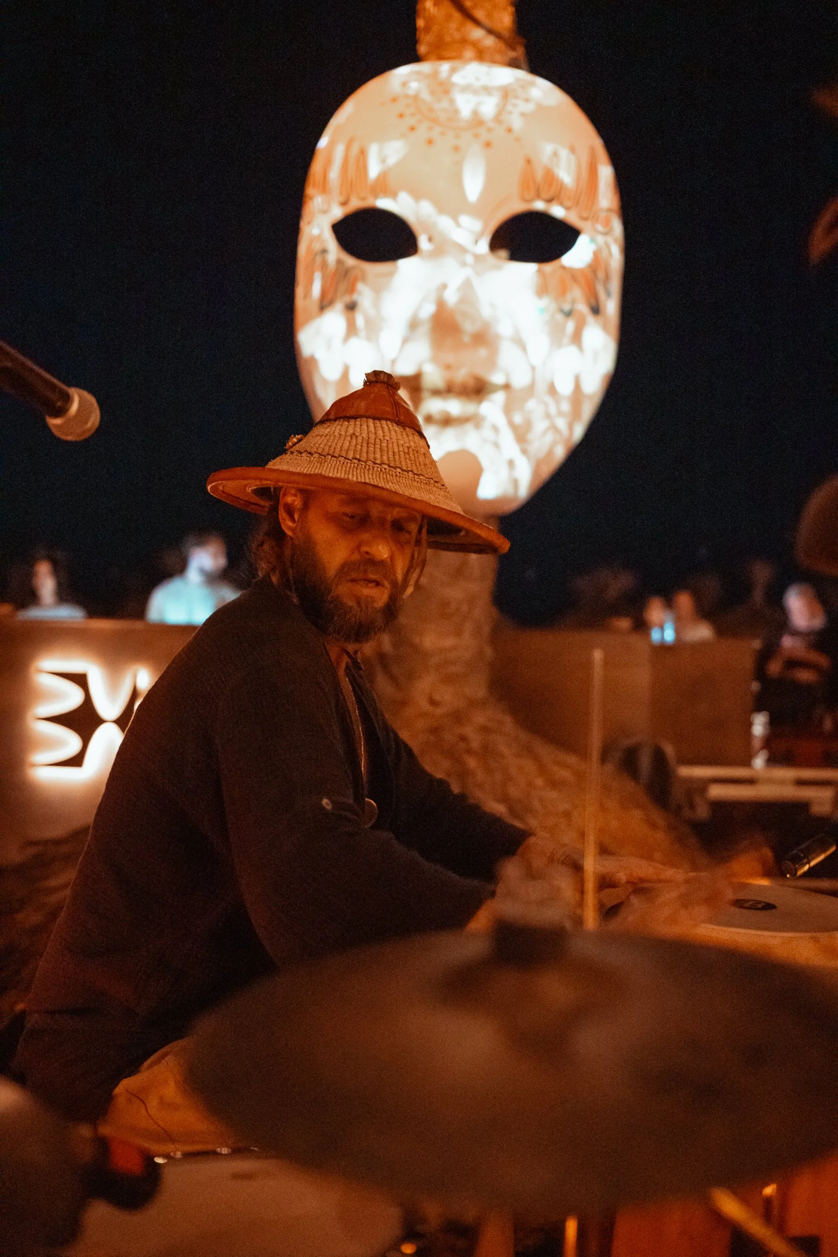 A man wearing a wide-brimmed straw hat sitting at a table outdoors at night, with a large illuminated skull projection in the background.