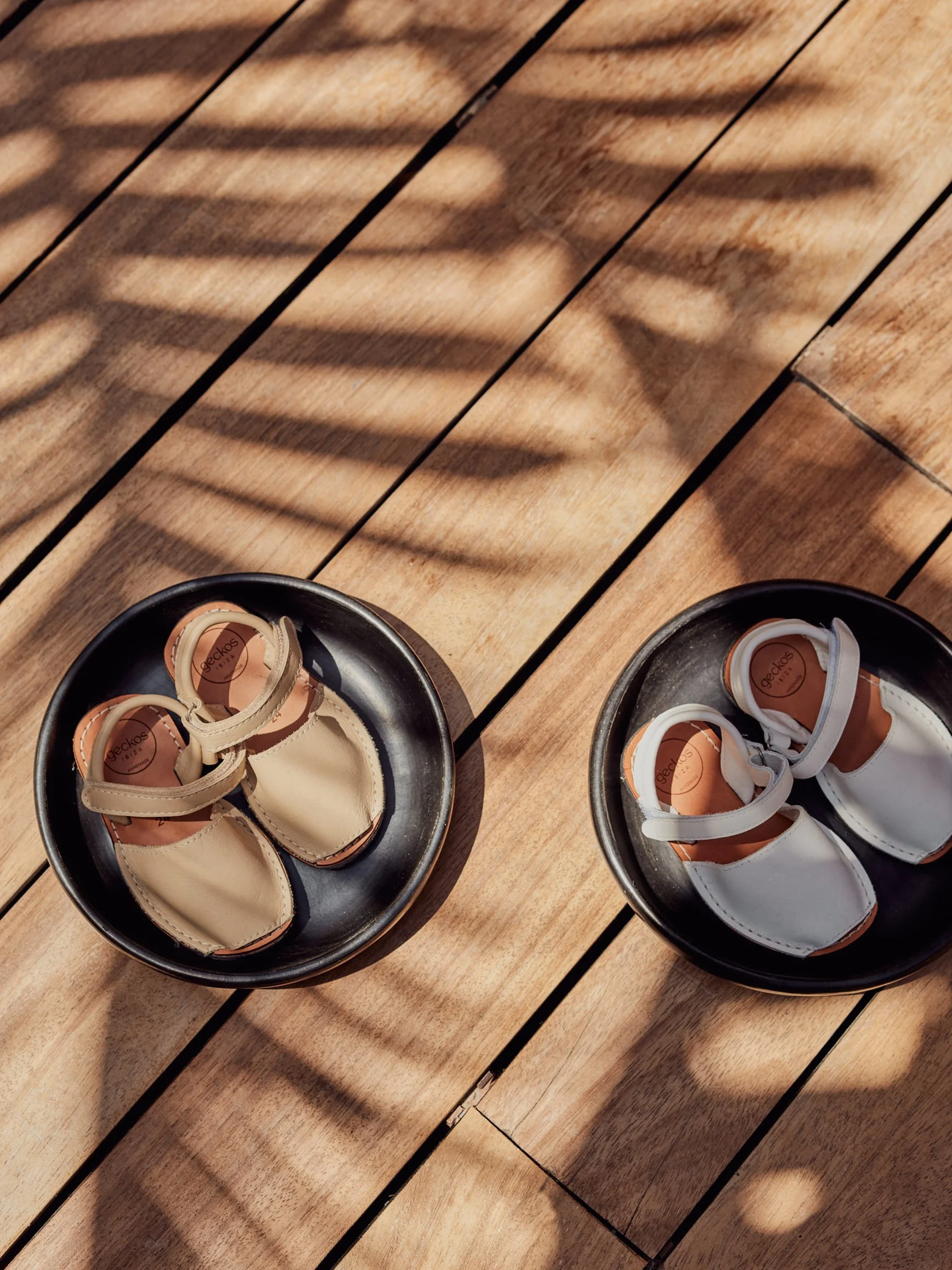 Pair of black trays, each holding a pair of small beige and white shoes, placed on a wooden surface with striped shadows.