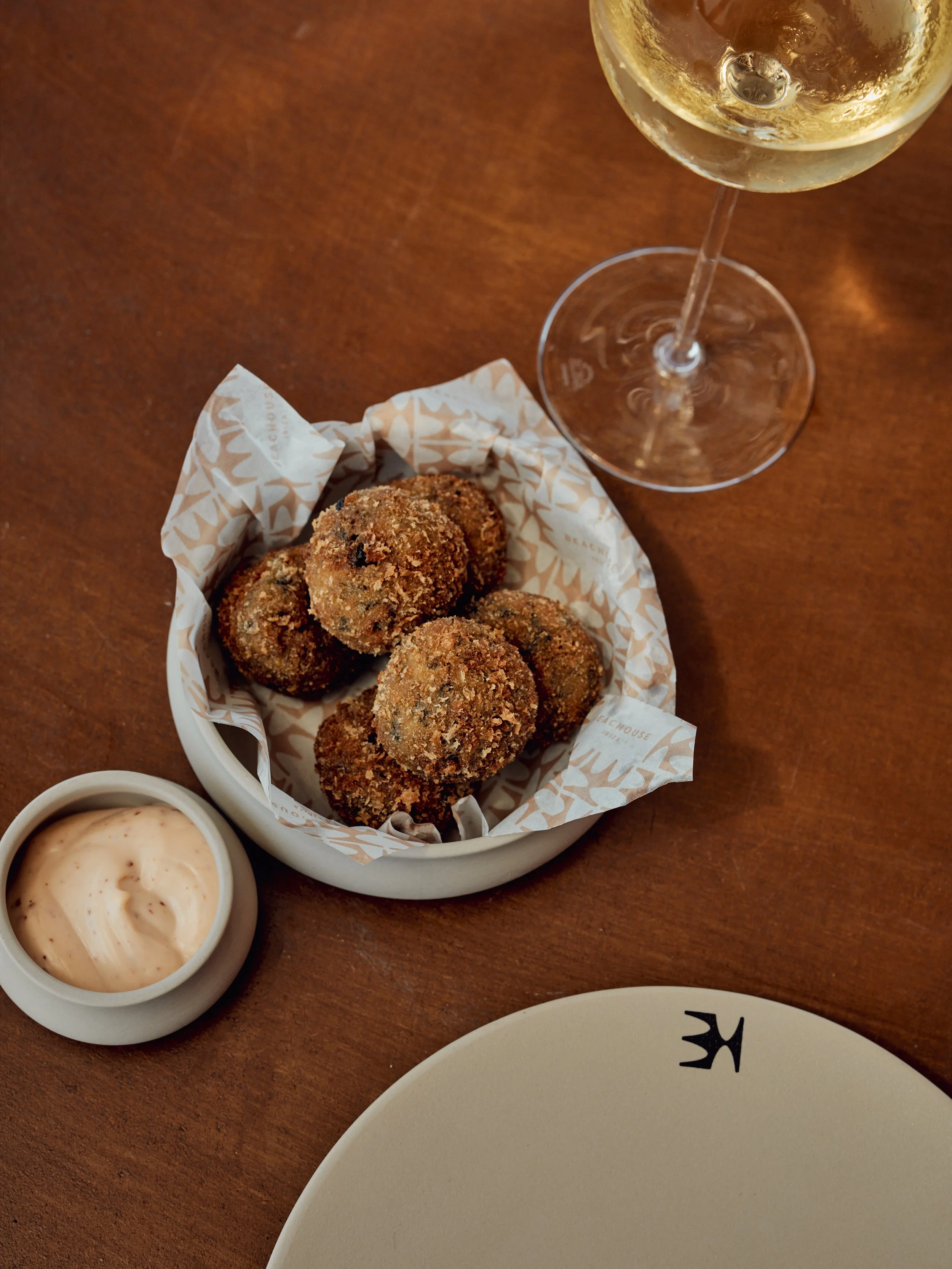 A basket of breaded fried snacks, a cup of dipping sauce, and a glass of white wine on a wooden table.