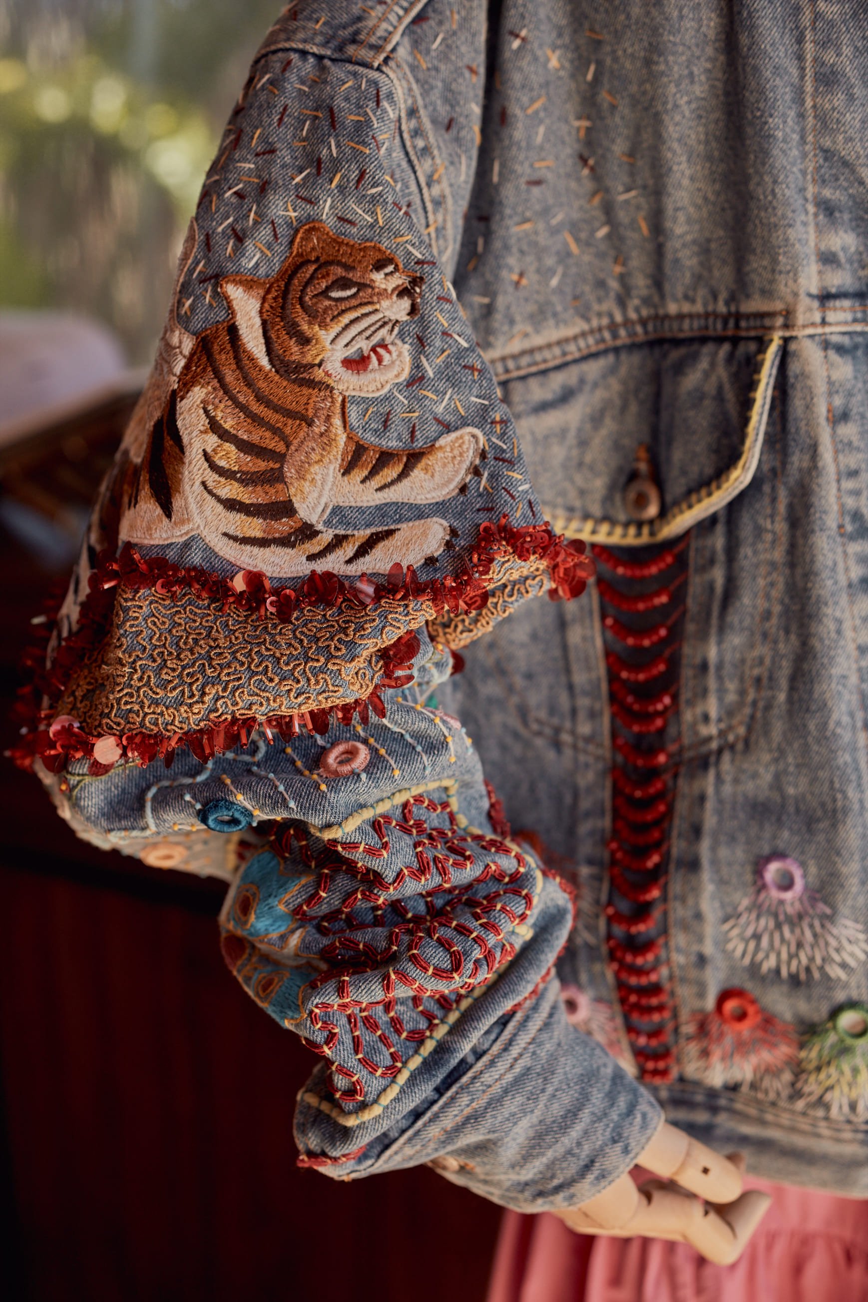Close-up of a denim jacket with colorful embroidery and patchwork details, including a tiger patch on the sleeve, with red beads on the cuffs, and part of a pink curtain in the background.
