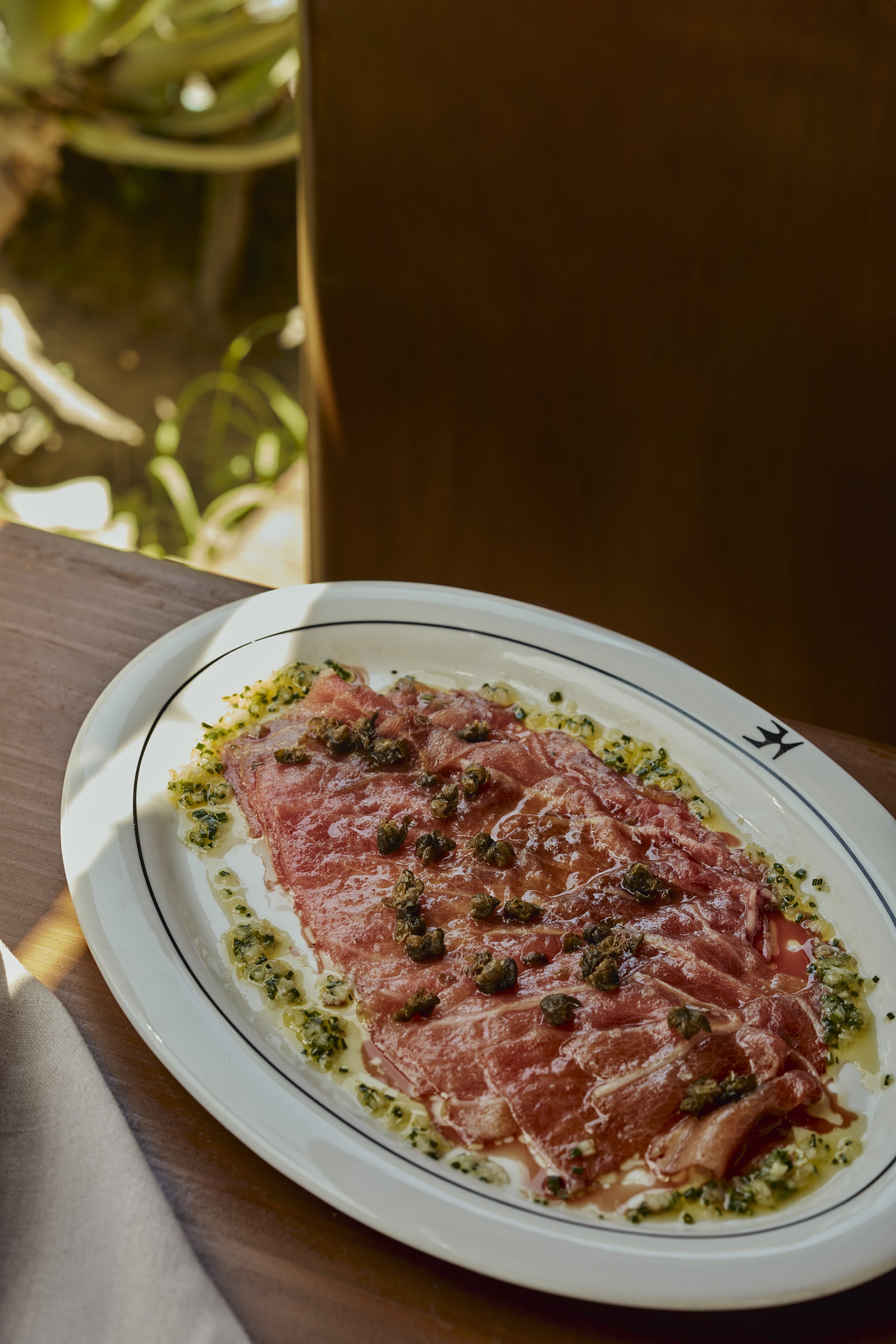 Thin slices of cured meat, possibly prosciutto, topped with black pepper, served on a white plate with a decorative border, garnished with chopped herbs and drizzled with oil.