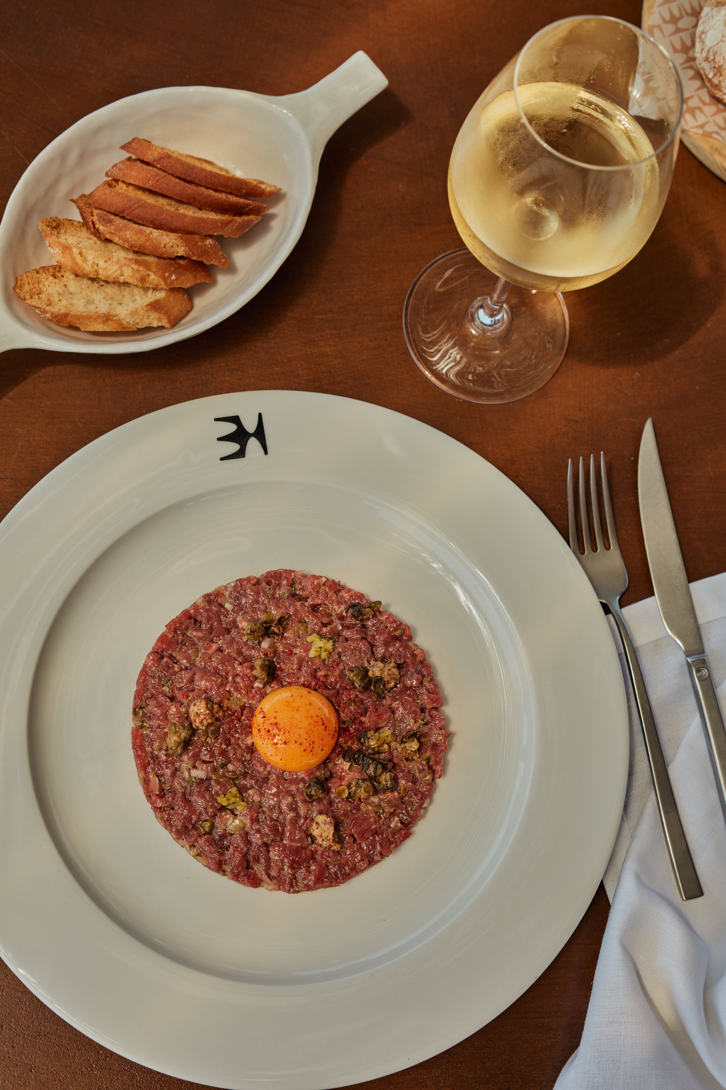 A plate of steak tartare topped with a raw egg yolk, a glass of white wine, and a basket of sliced bread on a wooden table.