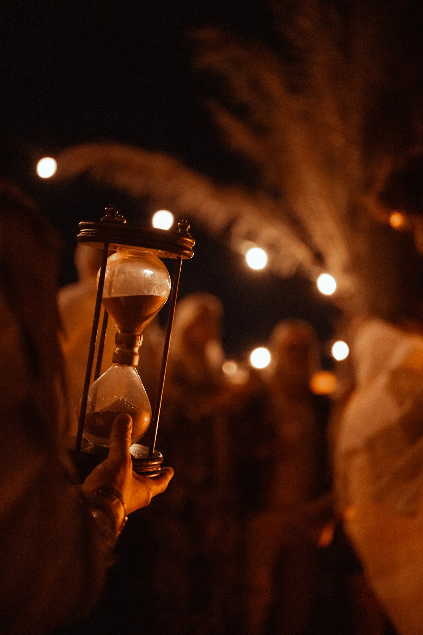 A person holding an hourglass illuminated by a lantern during a night event with string lights in the background.