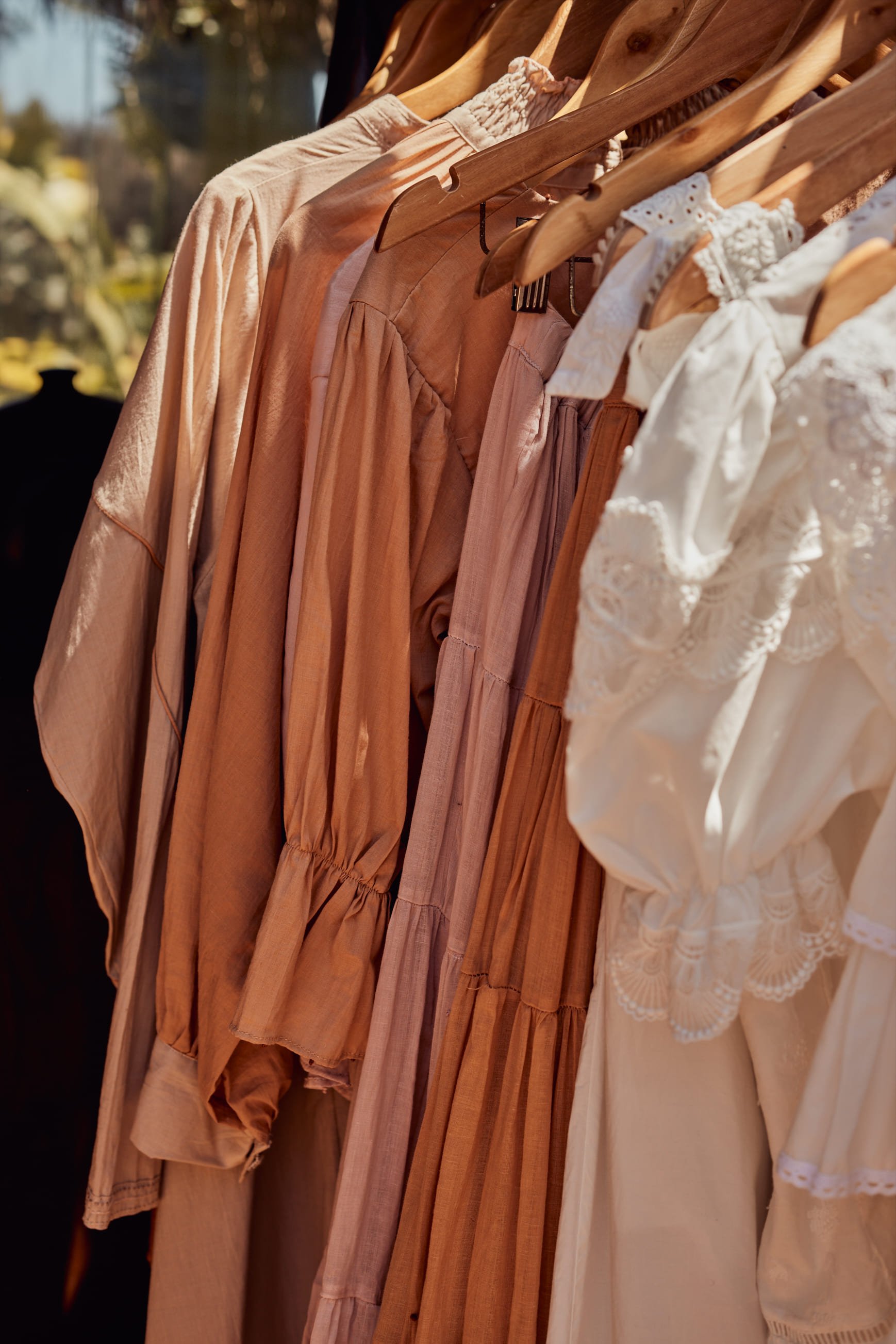 Various pastel-colored dresses and blouses hanging on a rack, with lace and ruffle details, in a softly lit setting.