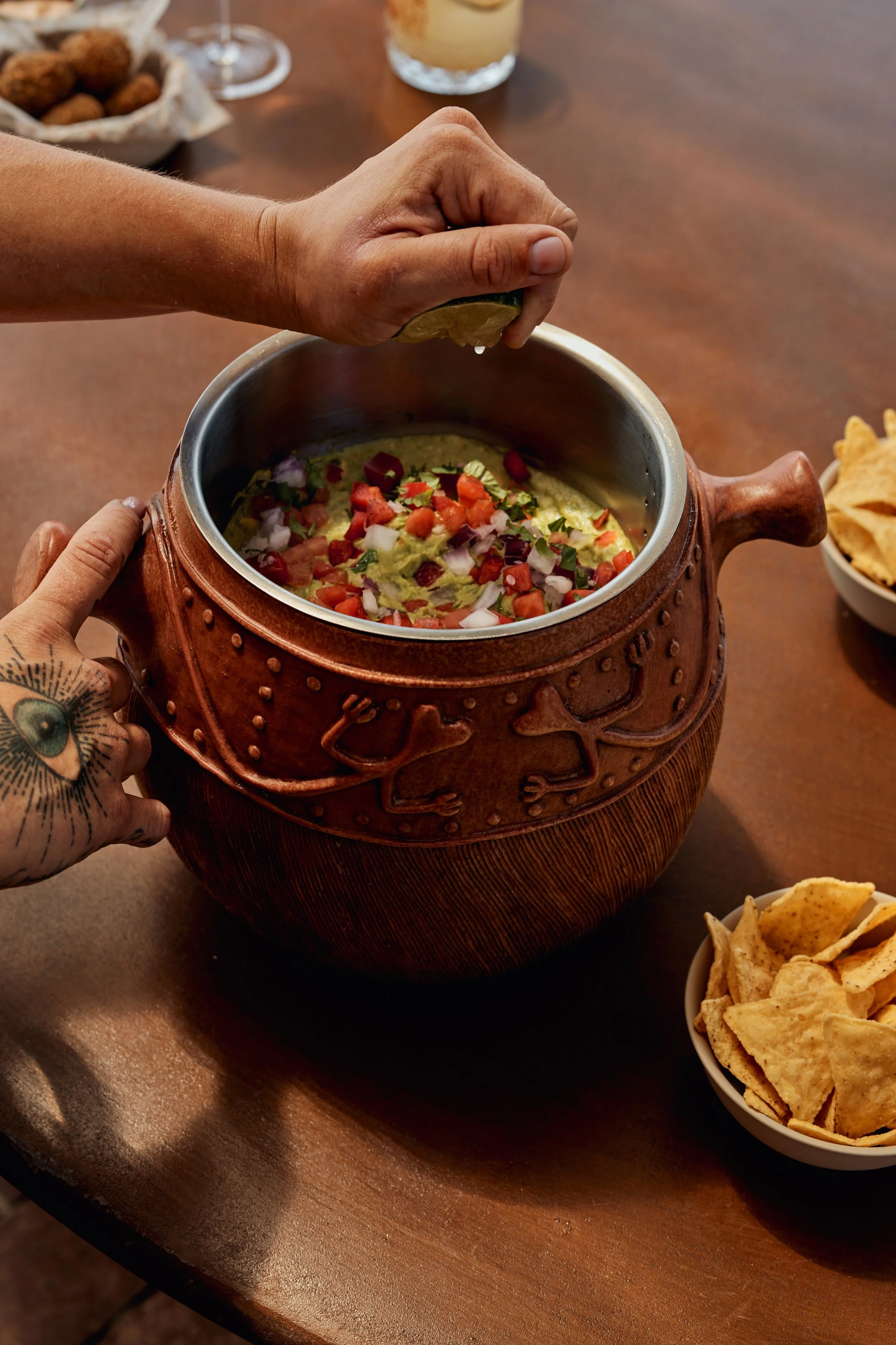 A hand squeezing lime into a pot of guacamole topped with chopped tomatoes, onions, and cilantro on a wooden table, with a bowl of chips nearby.