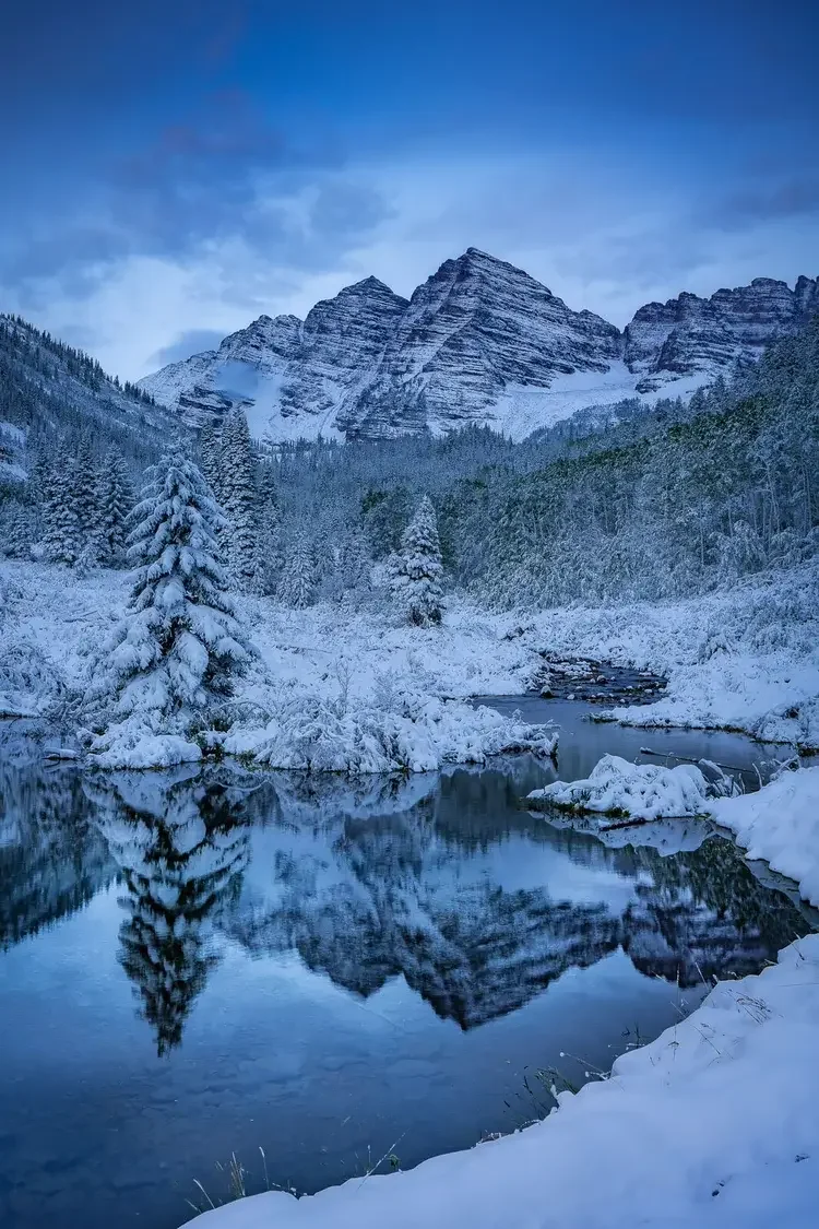Snow-covered trees and rocky mountains reflected in a calm river under a cloudy blue sky.
