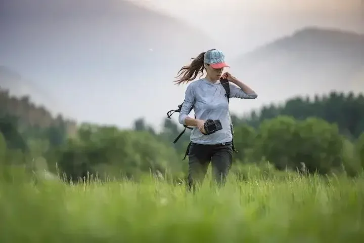 Tamara Susa walking through a grassy field, holding a camera, with mountains and trees in the background. Photo by Luke Tornare