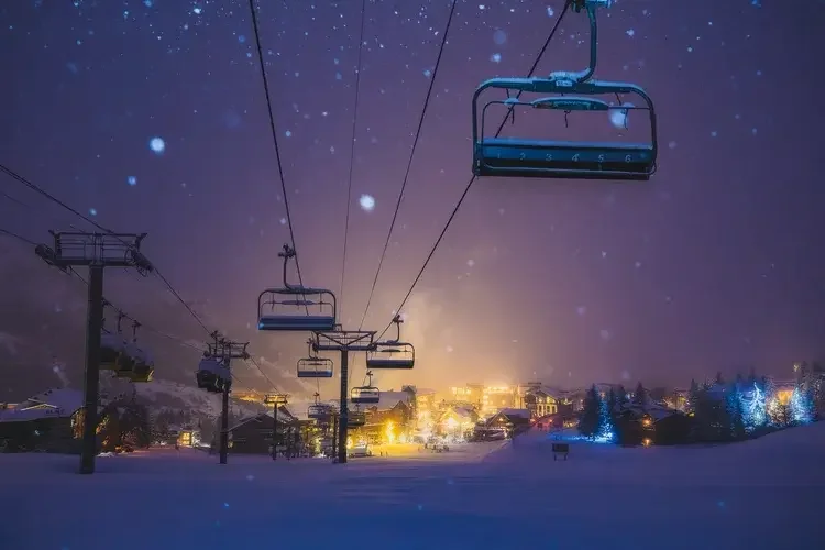 Nighttime view of snow-covered ski resort with empty ski lifts, illuminated buildings, and snow falling.
