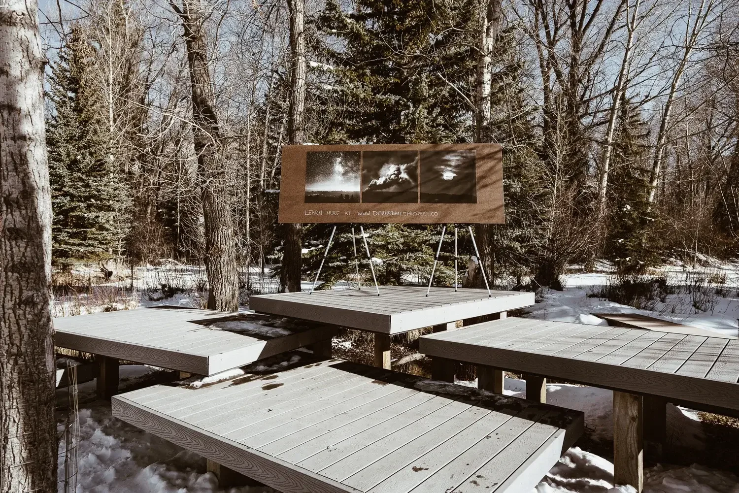 Outdoor wooden platform stage with a display board showing night sky and cloud images in a snowy forest.