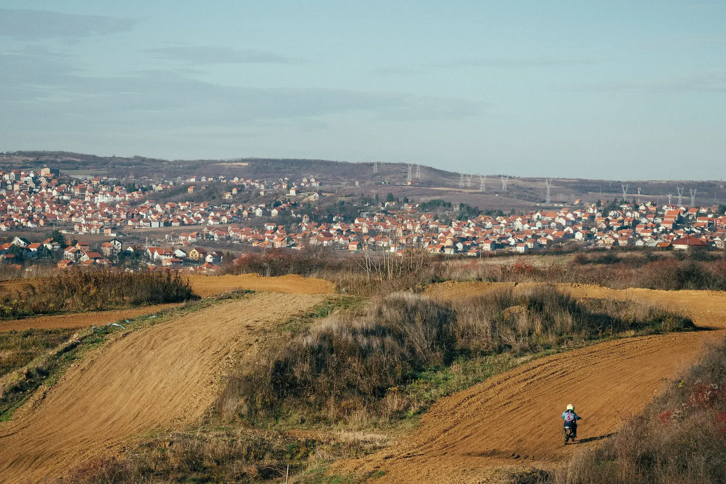 A dirt bike rider wearing a helmet riding on a dirt trail in a hilly landscape with a residential town in the background.
