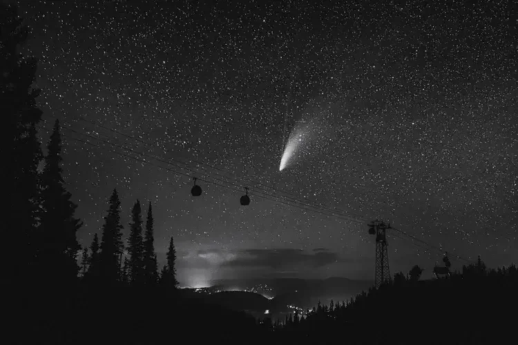 Nighttime landscape with star-filled sky, a bright comet, silhouettes of trees, a cable car system, and distant lights on the horizon.
