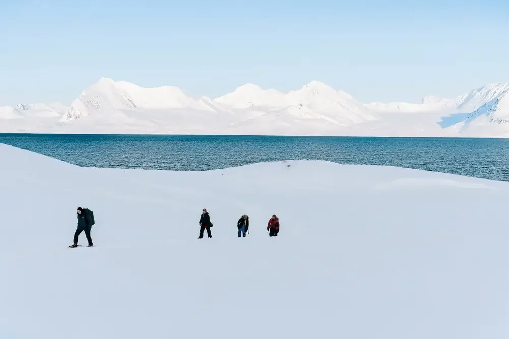 Four people dressed in winter gear walking through snow-covered landscape near a large body of water, with snow-capped mountains in the background.
