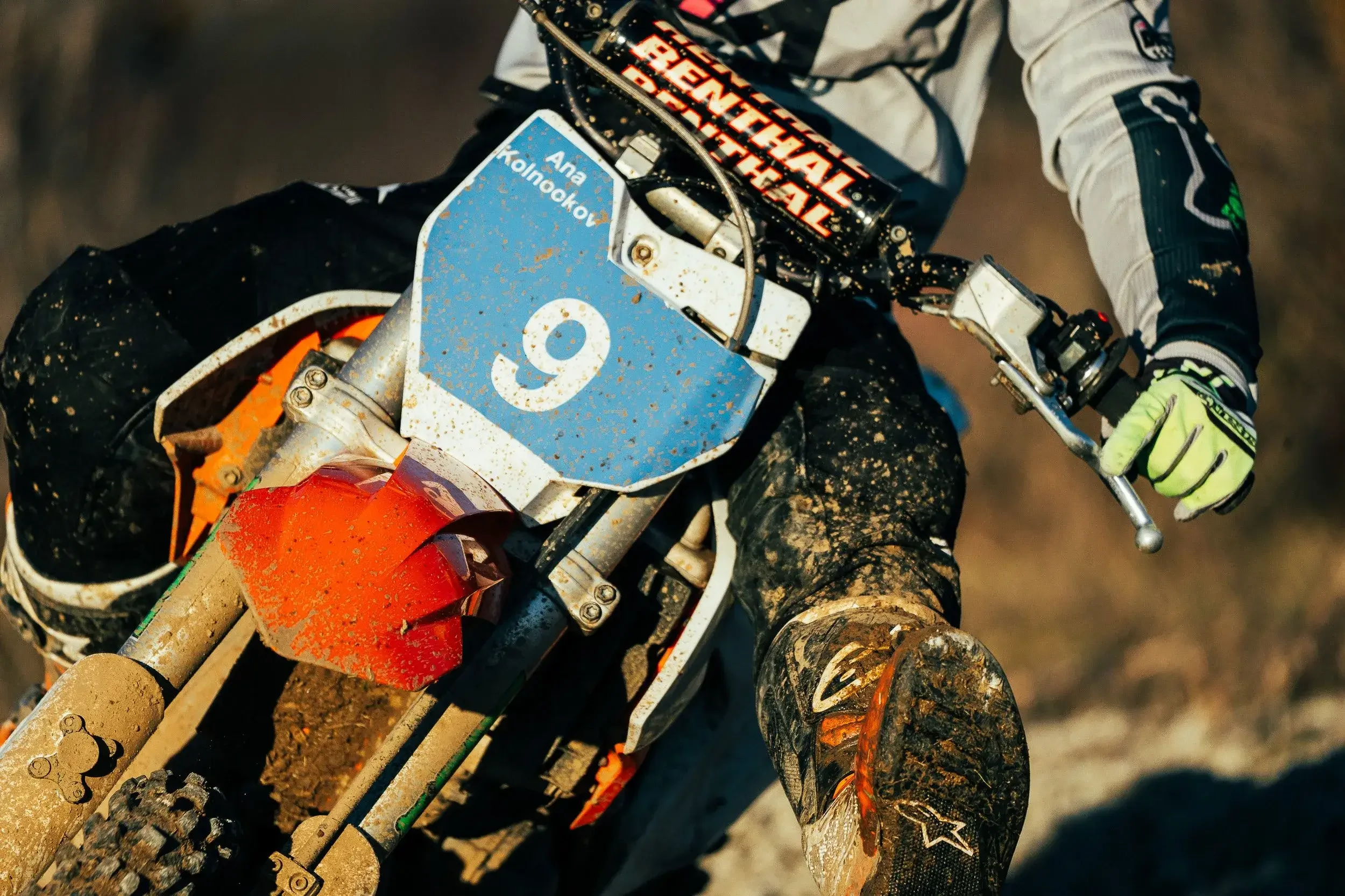 A motocross rider on a dirt bike wearing a white and black riding suit with yellow gloves, navigating through a muddy trail. The dirt bike has a blue number plate with the number 9 and mud splatters all over it.
