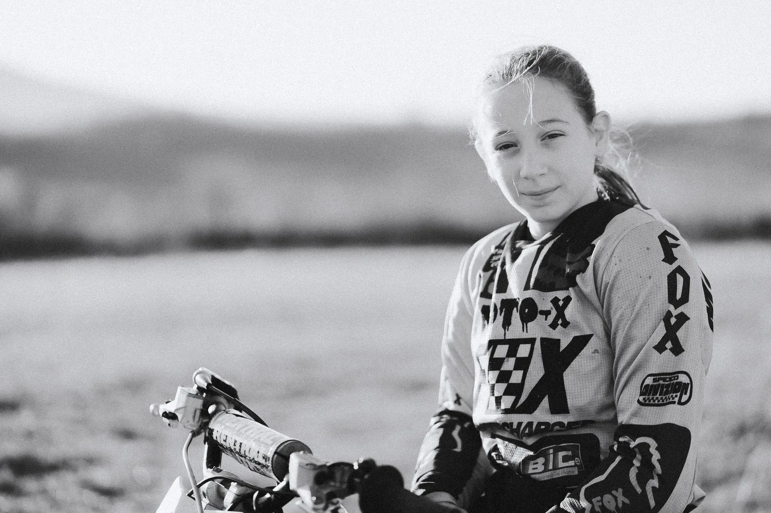 A young girl in motocross gear standing outdoors with a dirt bike, smiling at the camera.
