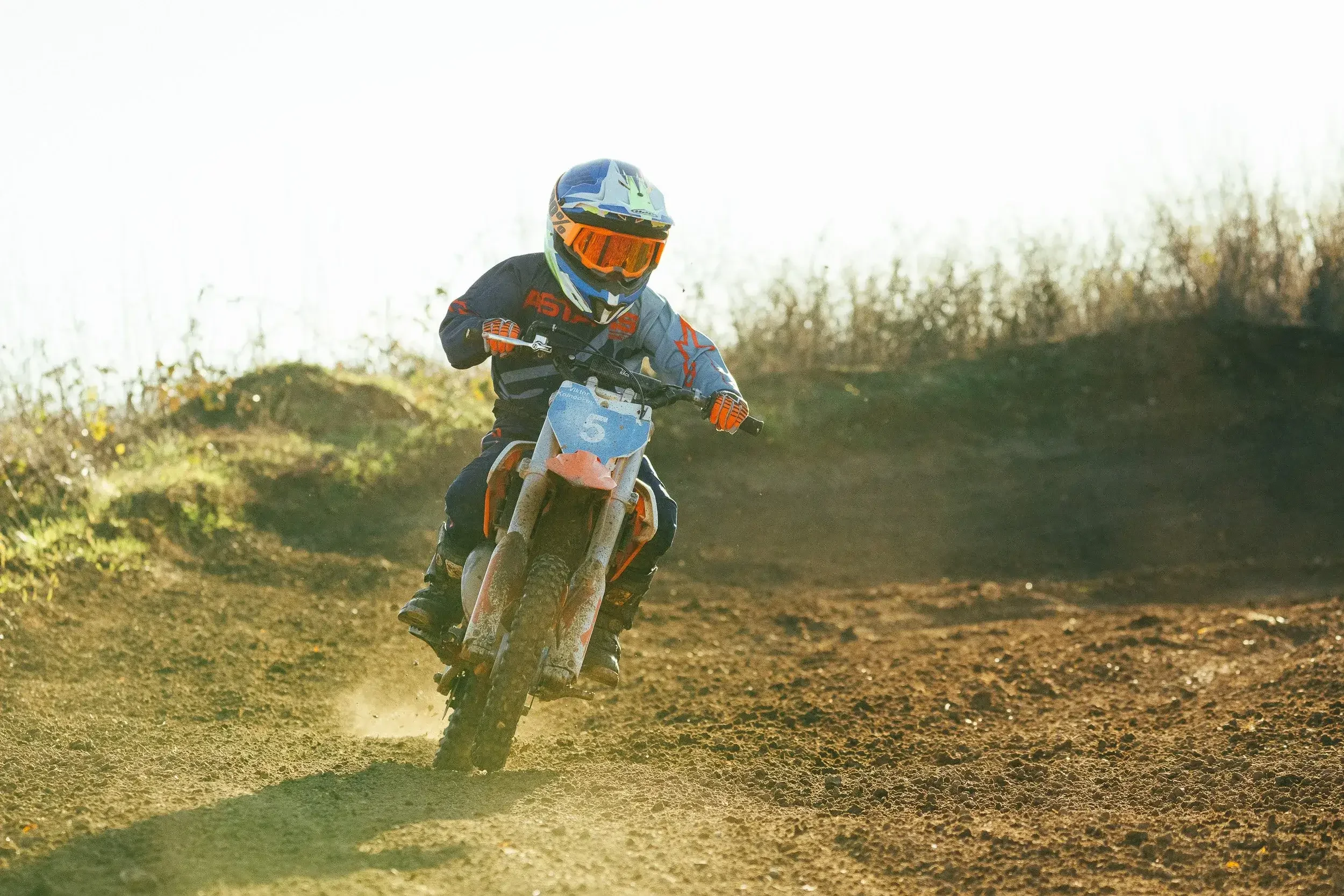 A young person riding a dirt bike on a rugged outdoor trail during daytime, wearing a helmet and protective gear.
