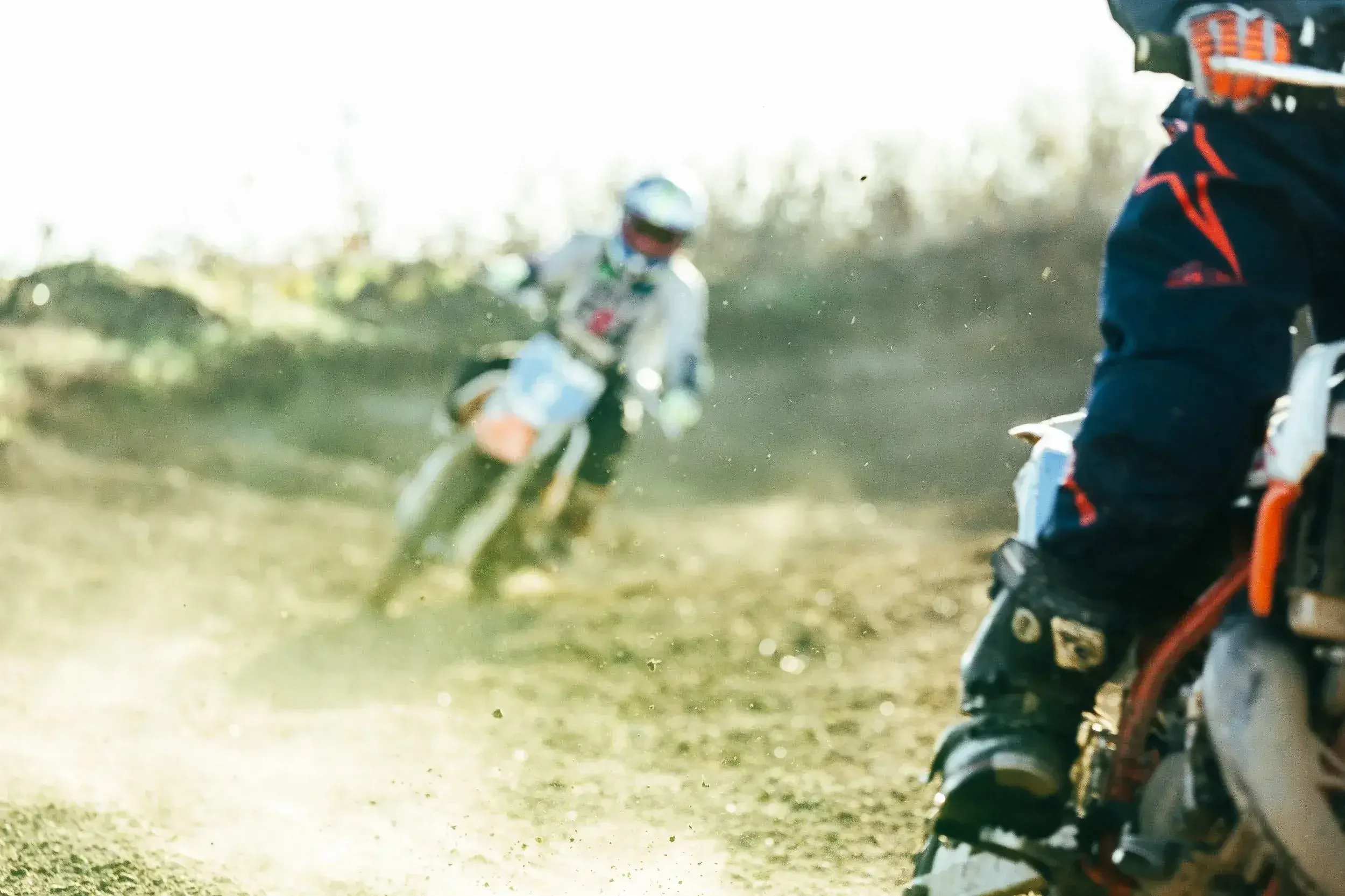 Two motocross riders in action on a dirt track, with one rider in focus on the right and a blurred rider in the background on the left.
