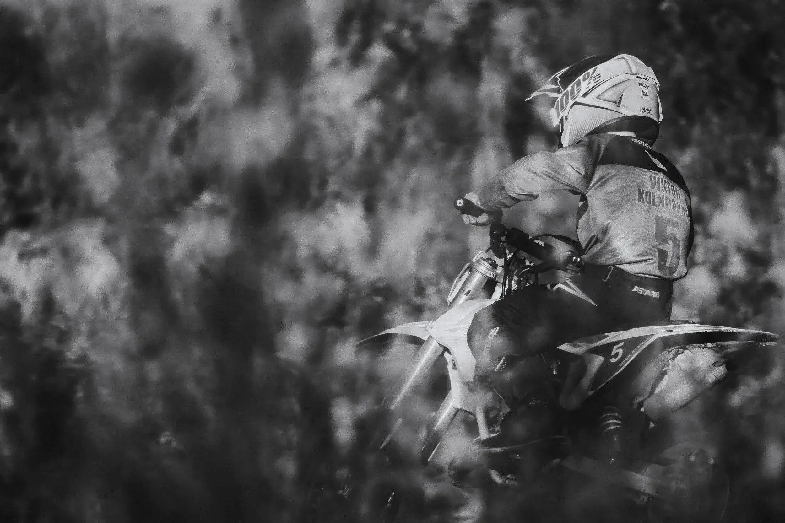 A person riding a dirt bike in an outdoor setting with a rocky background, wearing a helmet and protective gear, in black and white.
