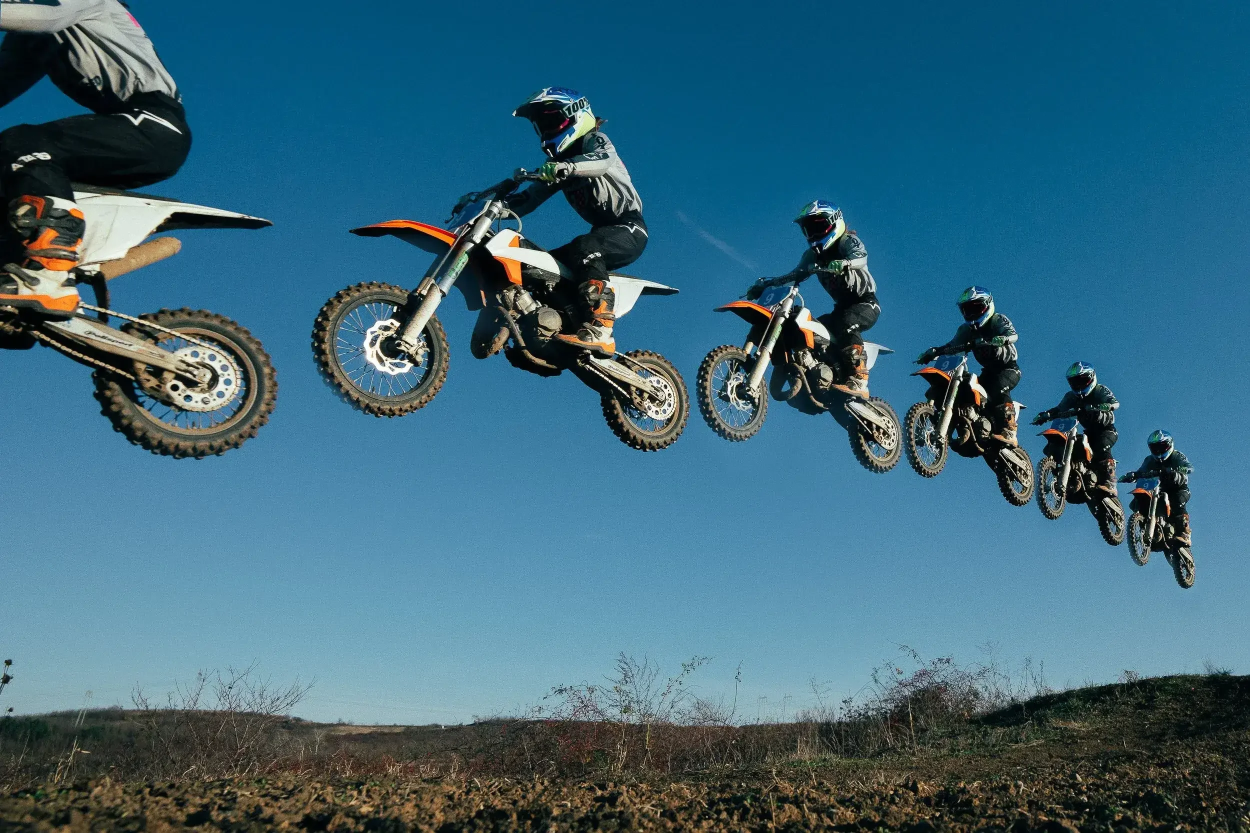 Multiple motocross riders in black and white gear and helmets, jumping in a line against a clear blue sky over a dirt track.
