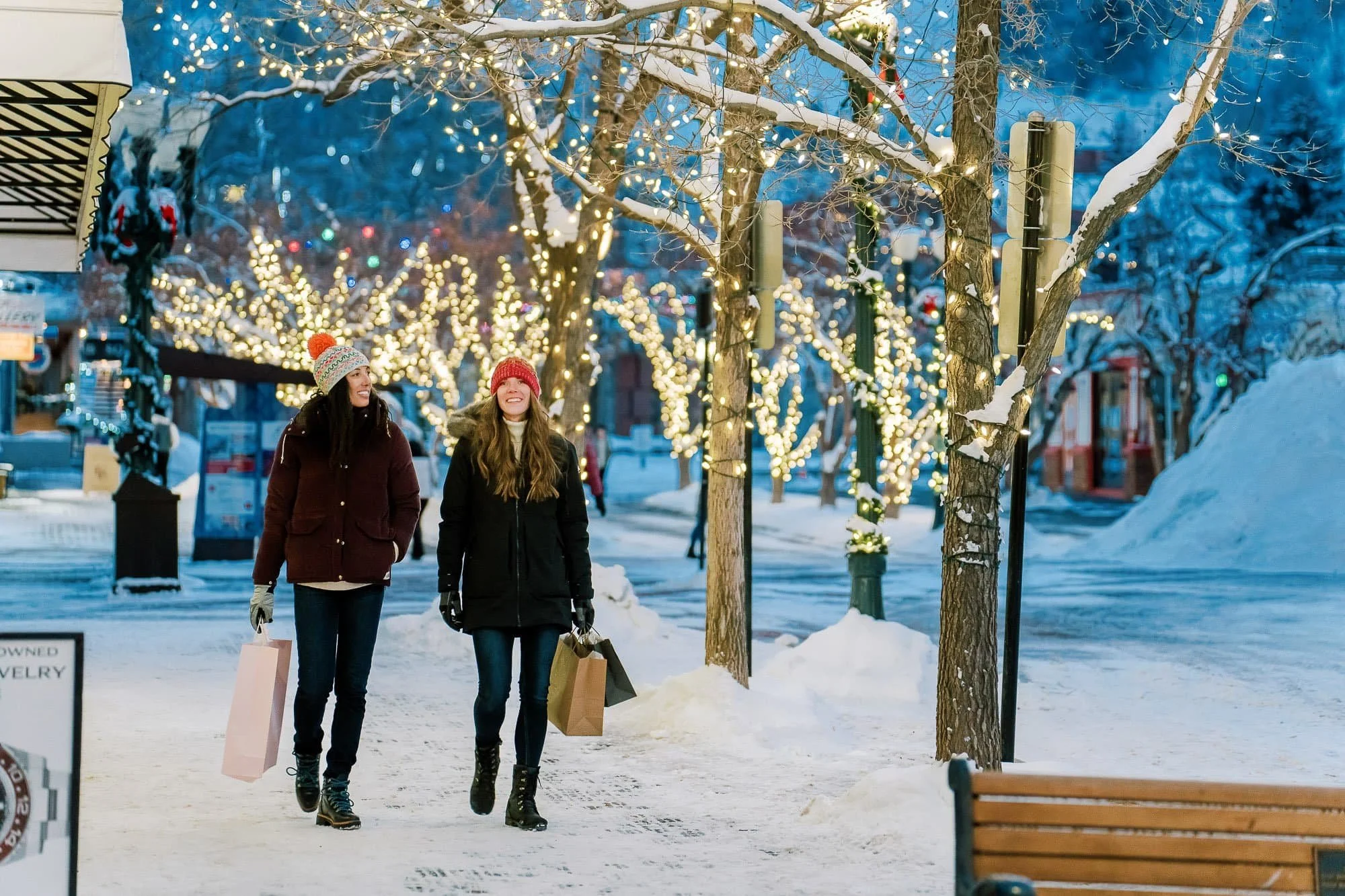 Two women walking and shopping outdoors on a snowy evening, decorated with Christmas lights on trees.