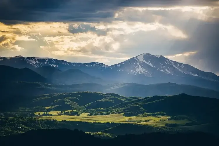 Scenic view of mountains with snow patches and green rolling hills under a cloudy sky with sunlight breaking through.
